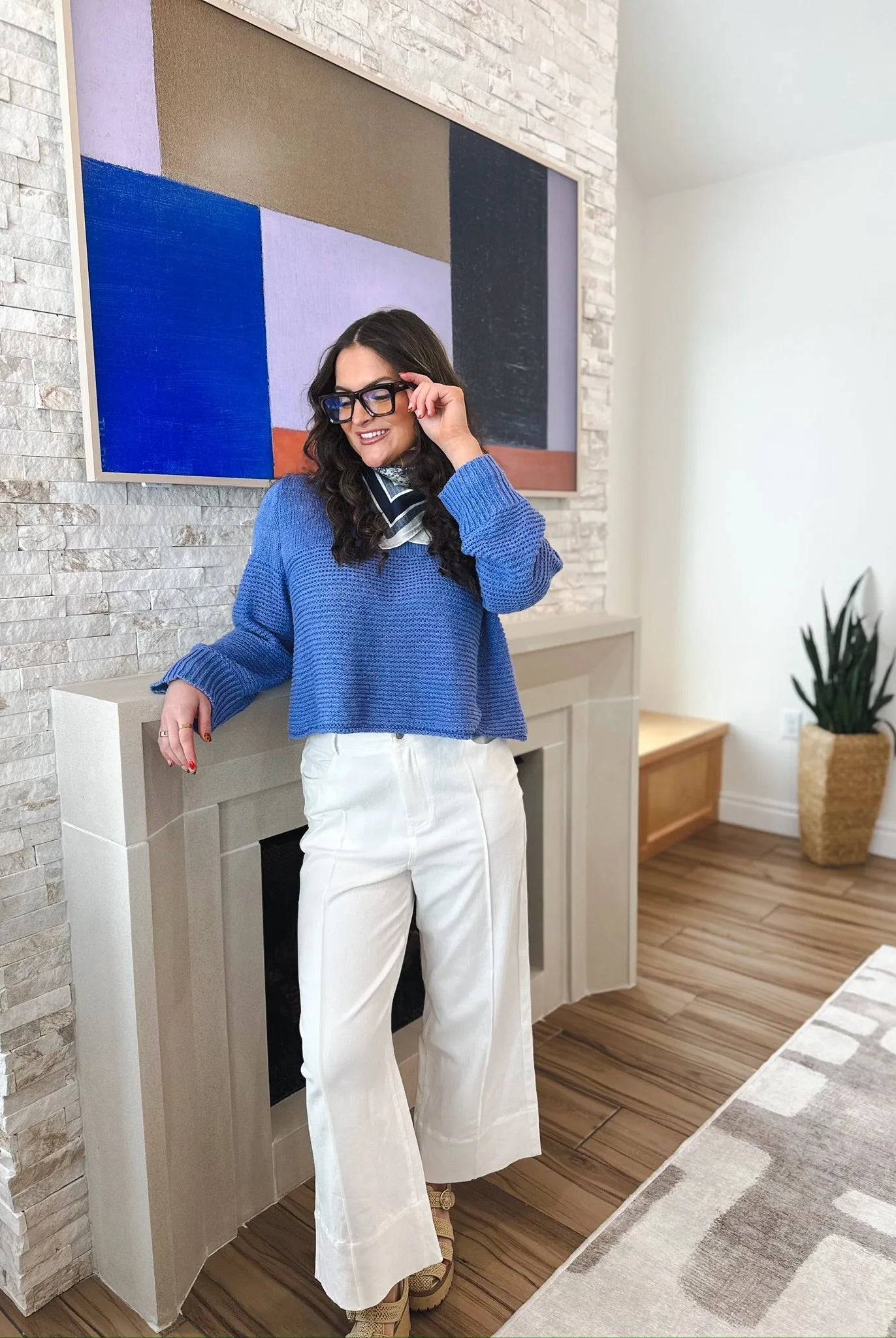 Woman wearing white wide-leg pleated denim jeans and a blue sweater, standing by a fireplace.