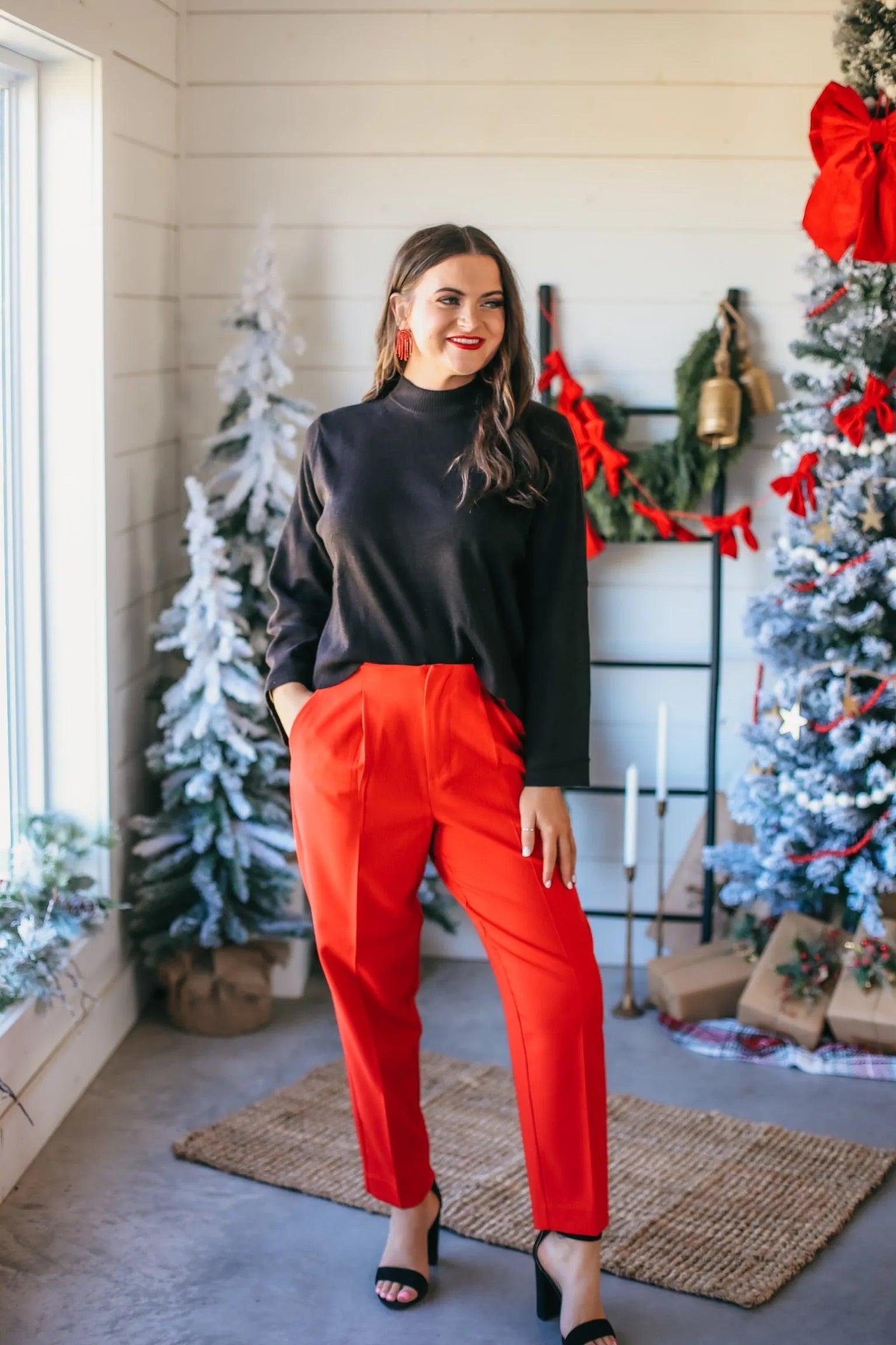 Entro woman in a black mock neck wide sleeve sweater and red high-waisted pants, standing in a festive holiday setting.