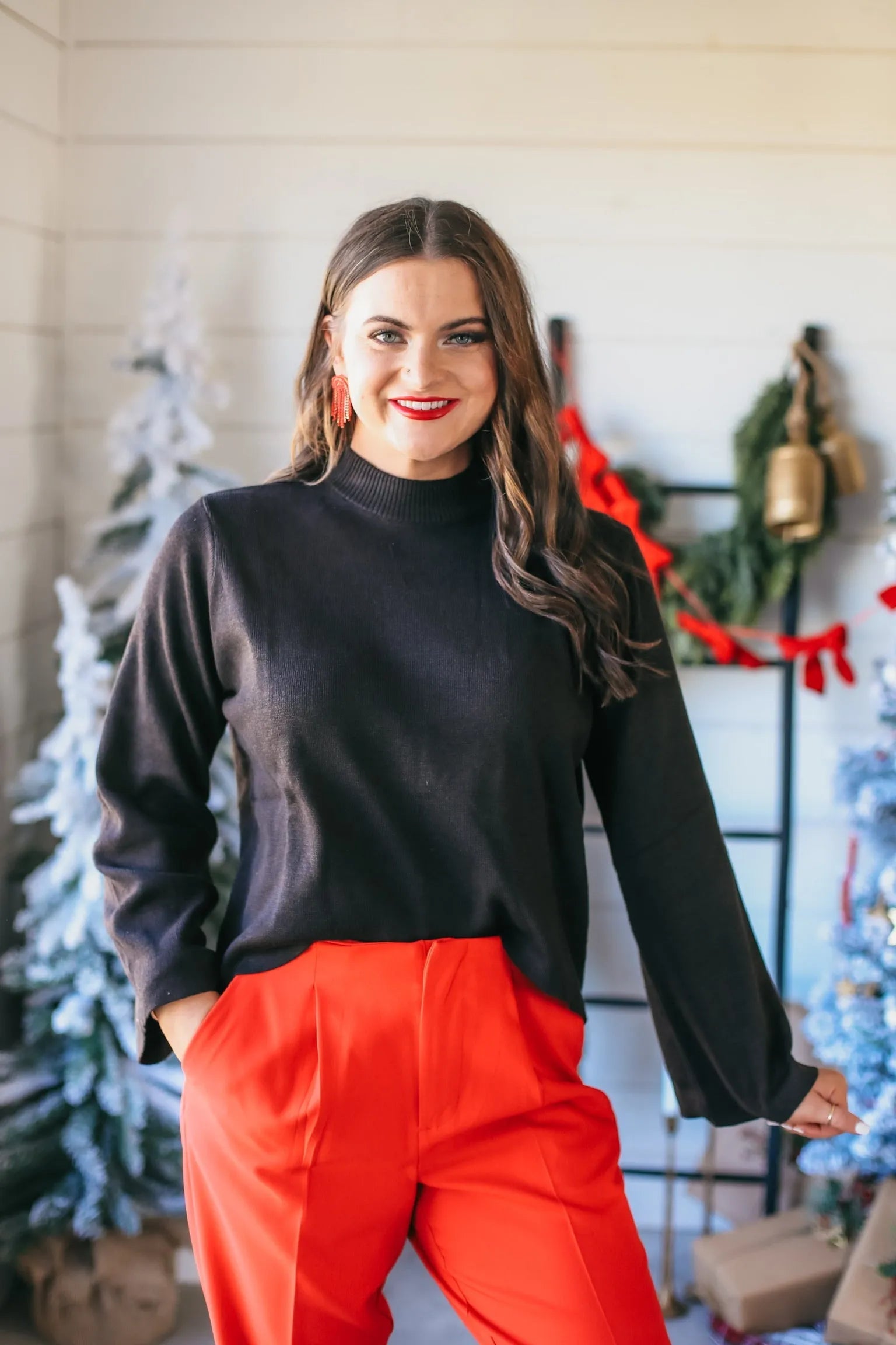 Woman wearing a black wide-sleeve mock neck sweater and bright red high-waisted trousers. She has on red earrings and red