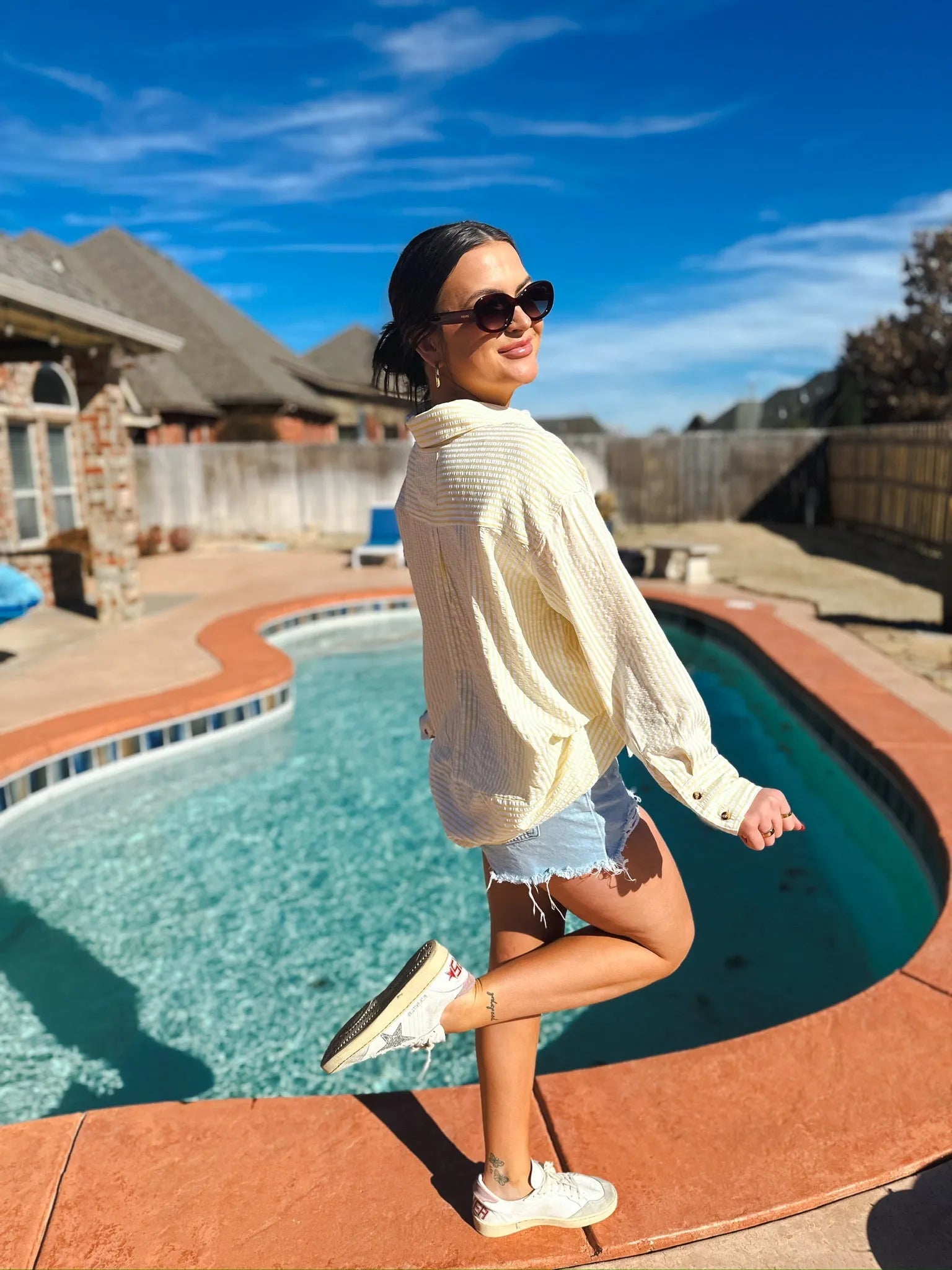 Woman wearing a yellow and white striped button-down shirt and denim shorts, standing by a pool. Sunglasses on.