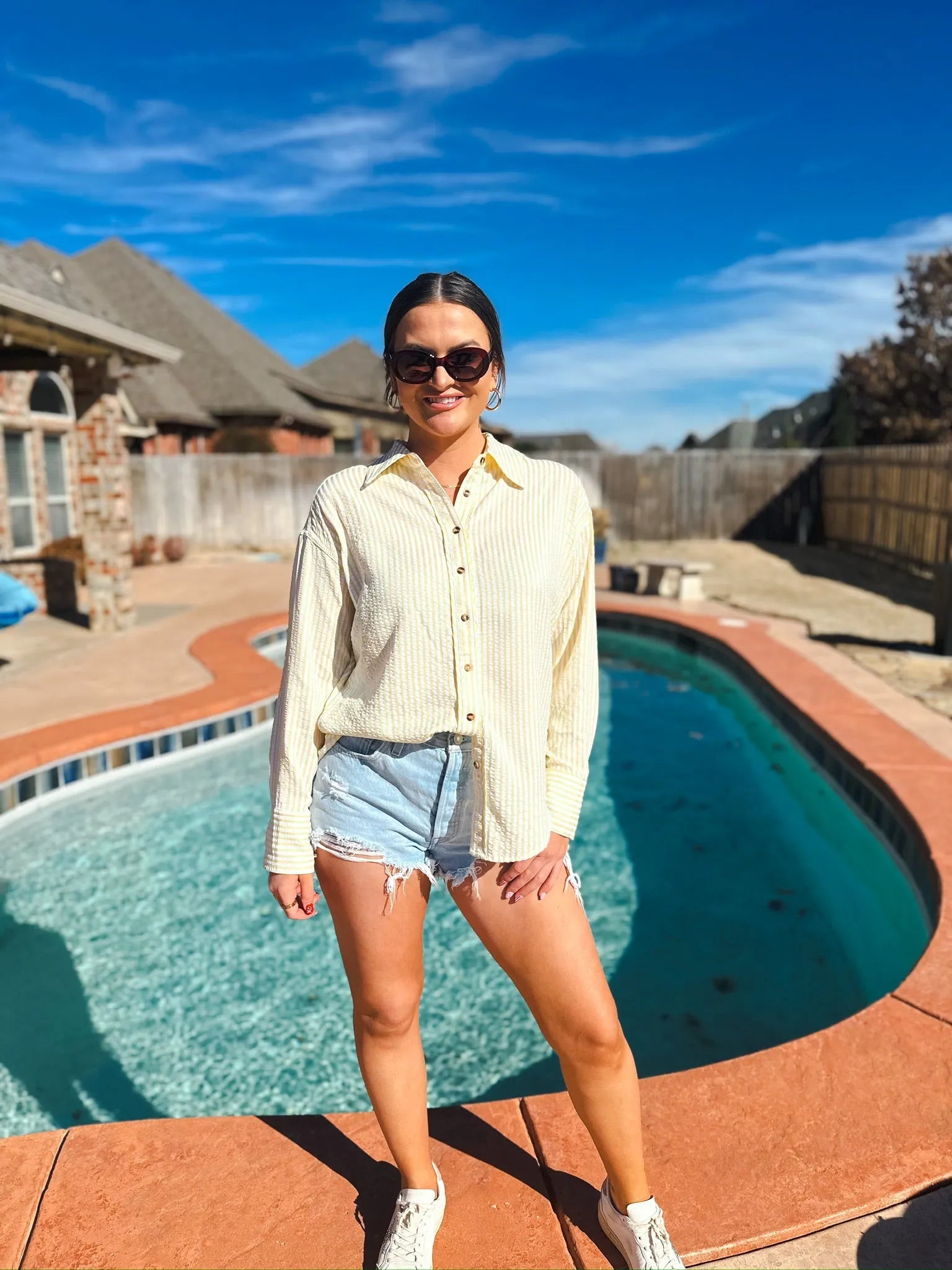 Woman wearing a yellow and white striped button-down shirt and distressed denim shorts, standing by a pool.