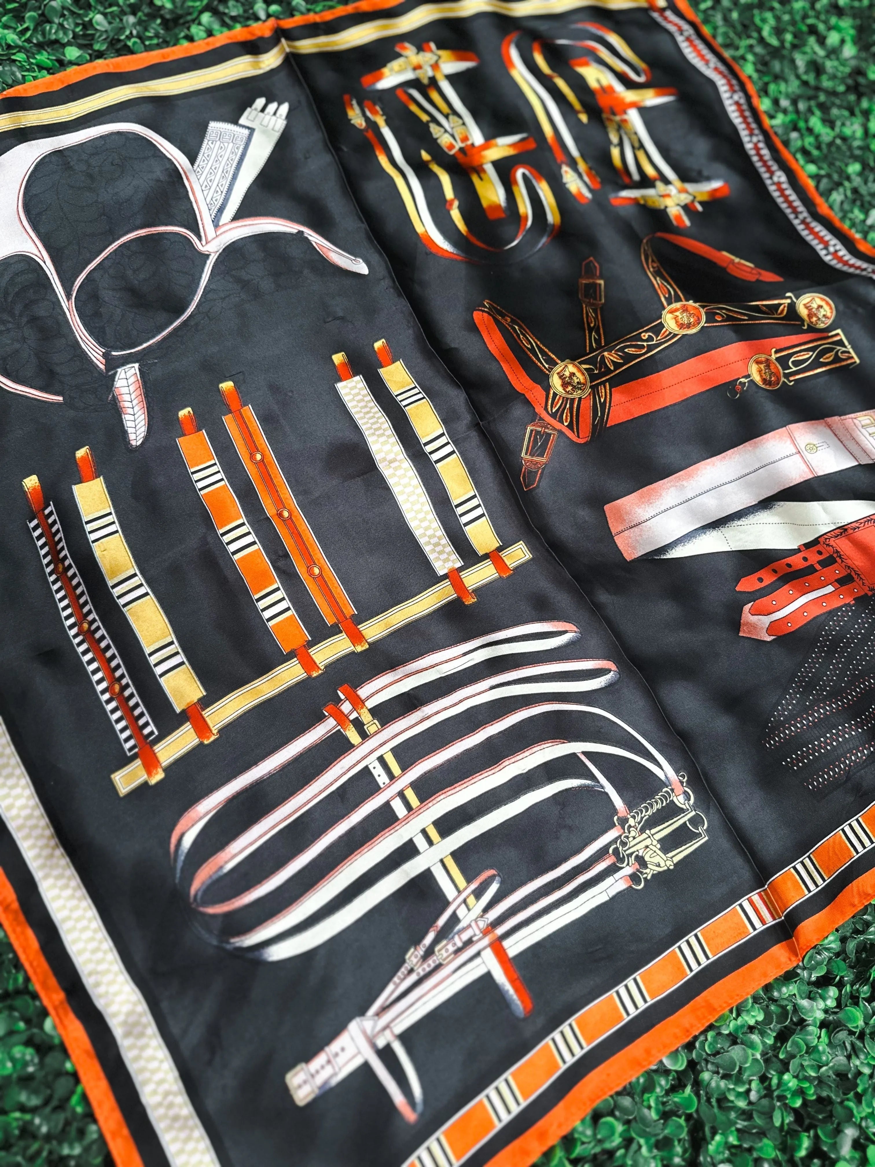 Black silk scarf with equestrian print: stirrups, bridles, and reins in white, orange, and gold accents. Features a