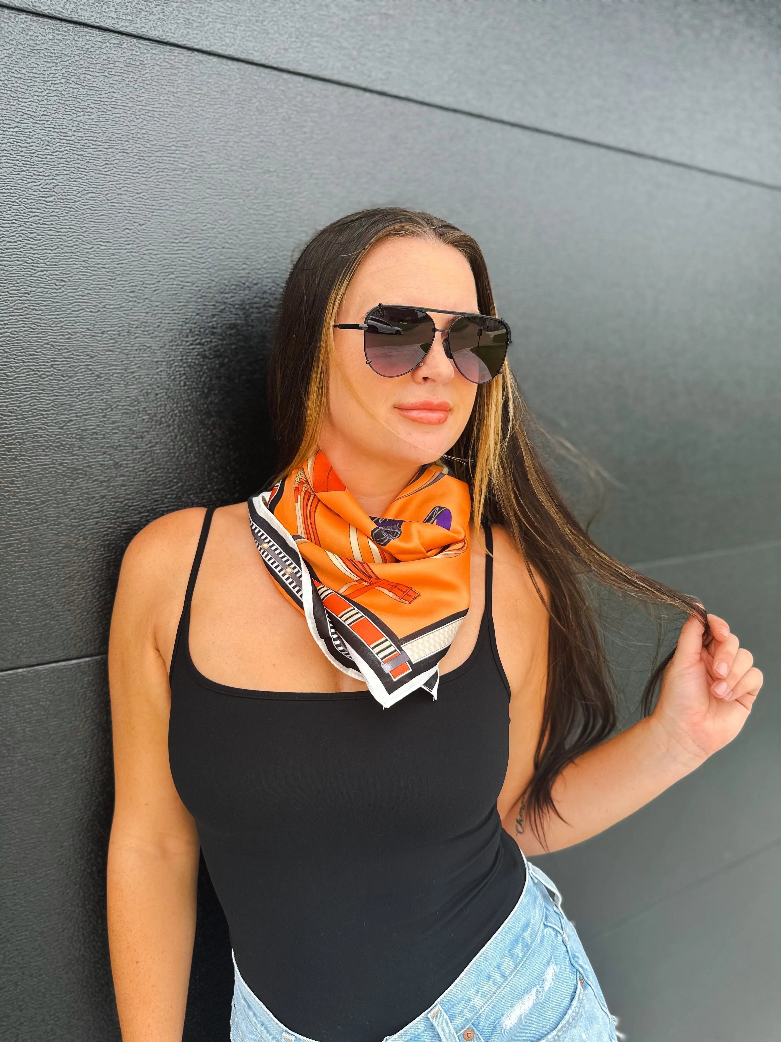 Model wearing an orange silk-texture square scarf with equestrian print. The scarf has black and white geometric borders