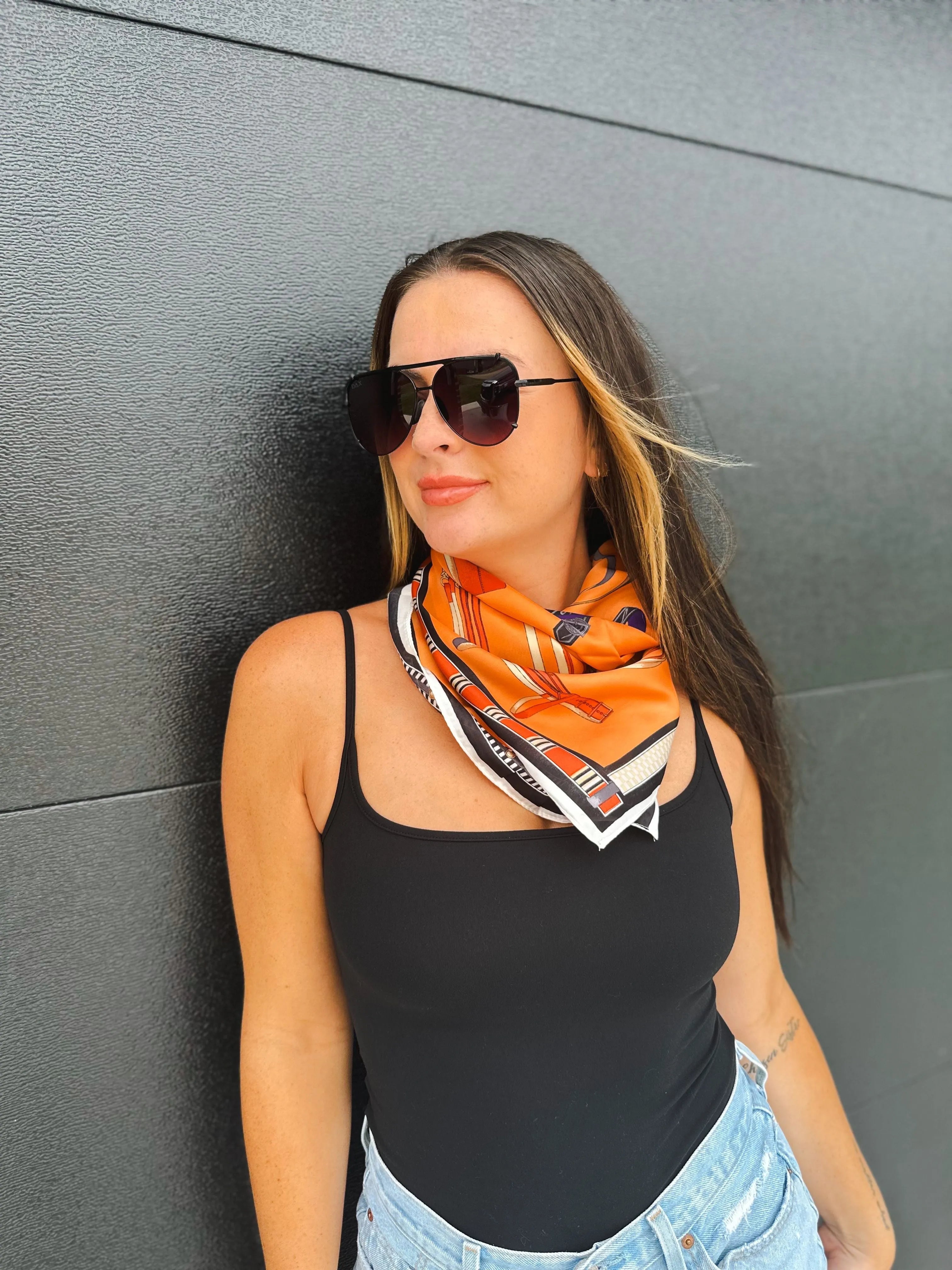 Woman wearing a black tank top and blue jeans, with an orange silk scarf featuring an equestrian print around her neck. She