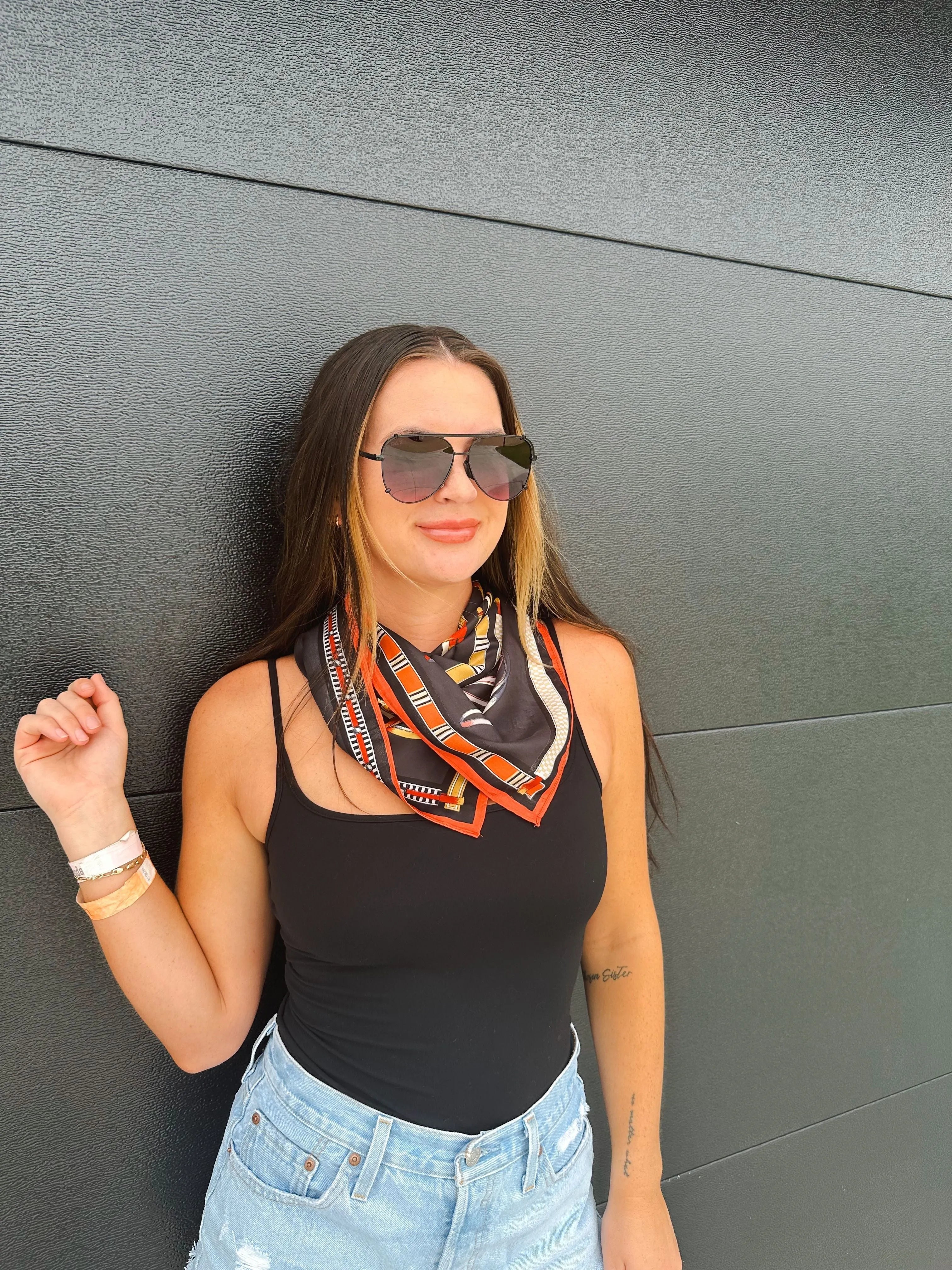 Woman wearing a black tank top, denim shorts, and sunglasses, with a square silk scarf featuring an equestrian print with