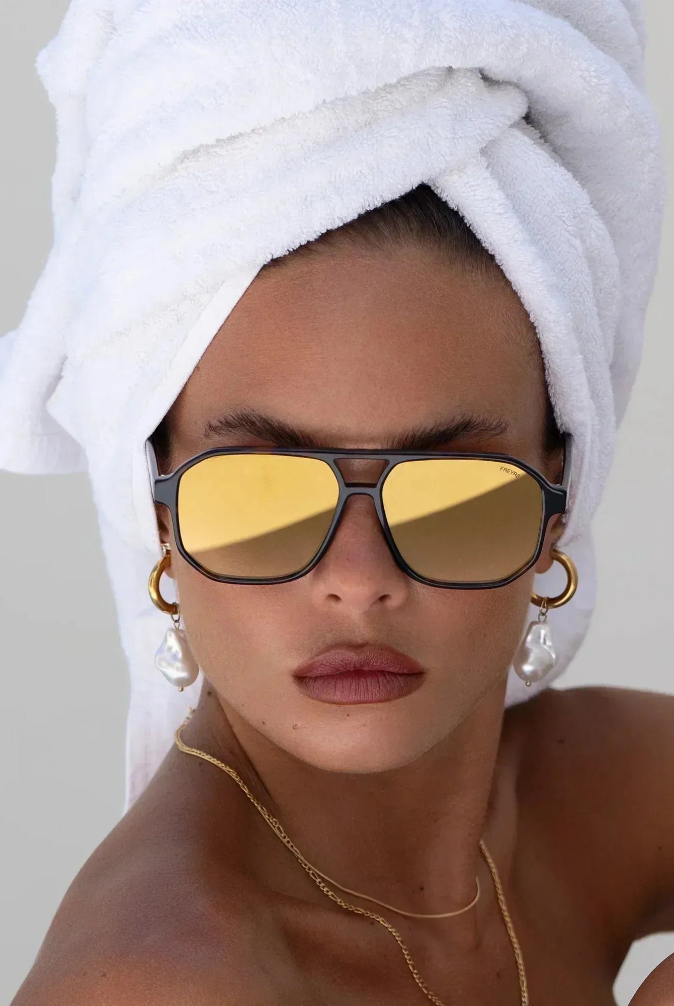 Freyrs Billie unisex aviator sunglasses with gold mirrored lenses and dark frames. A woman wears them with a white towel