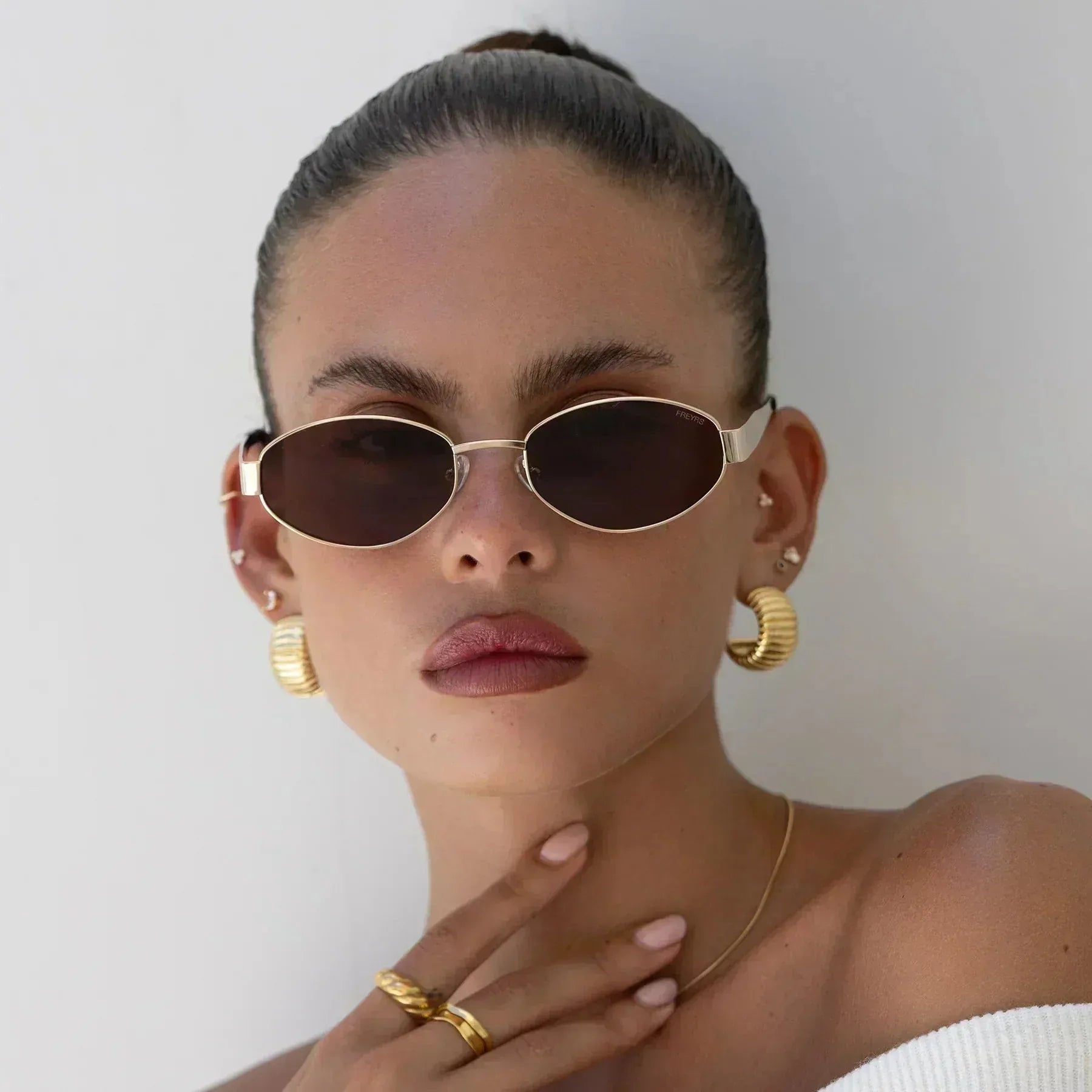 Freyrs Soho small oval sunglasses with gold rims and dark lenses, worn by a model with textured gold hoop earrings and rings.