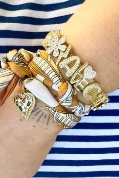 Gold Gemelli slider bracelet with charms, including a heart with colorful gems and 'IAM' text, worn on a wrist with a