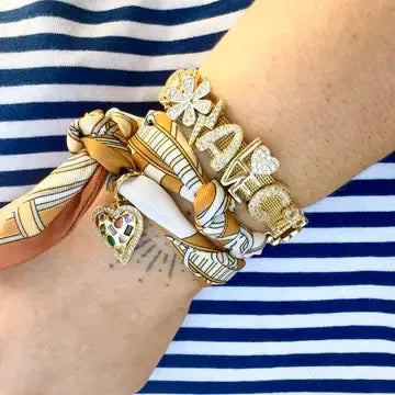 Gold Gemelli slider bracelet with charms, including a heart with colorful gems and 'IAM' text, worn on a wrist with a