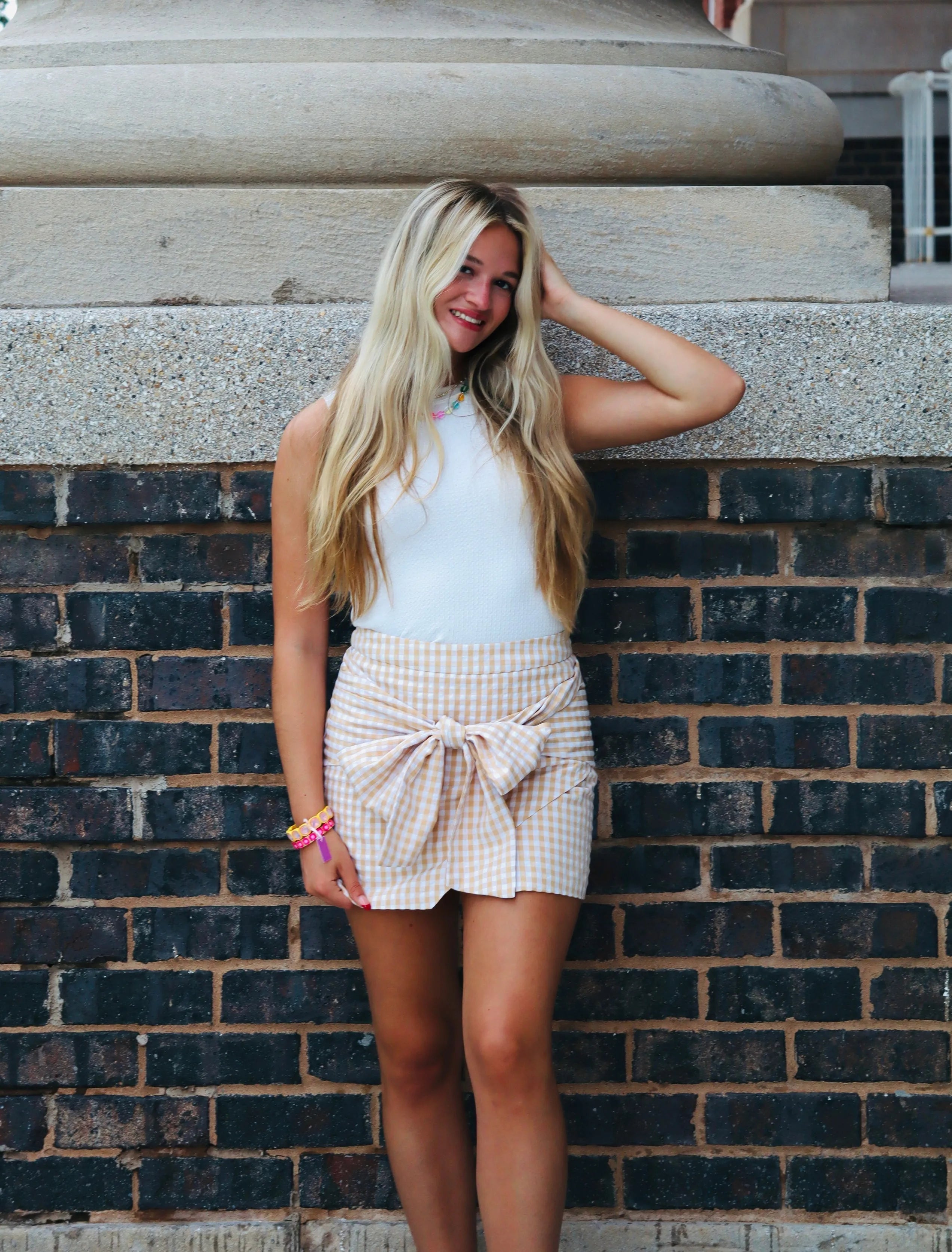 Woman in white top and beige gingham pencil skirt with a large front tie, standing against a brick wall.