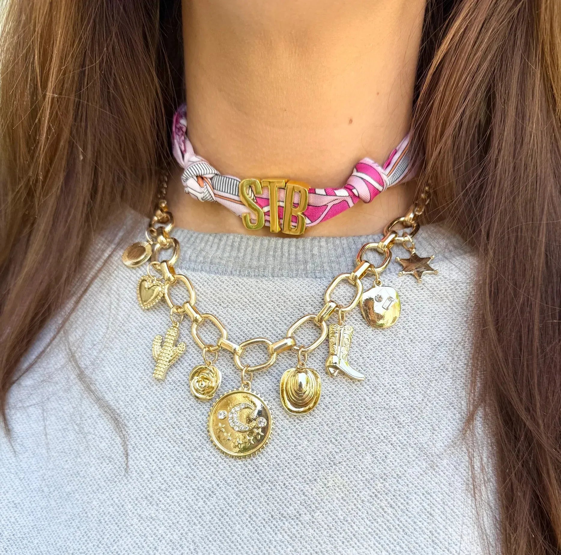 Gold initial charm necklace with "STB" letters, featuring charms like a cowboy boot, cactus, and moon on a gold chain, worn