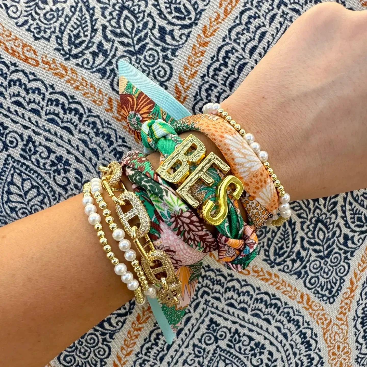 Gold initial charms spelling "BES" on colorful patterned bracelets worn on a wrist, alongside pearl and gold bead bracelets.