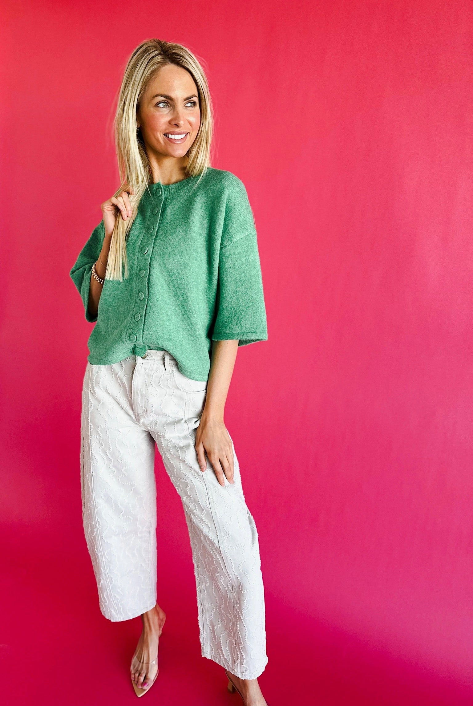 Woman wearing a green short sleeve sweater cardigan and white patterned wide leg pants against a bright pink backdrop.