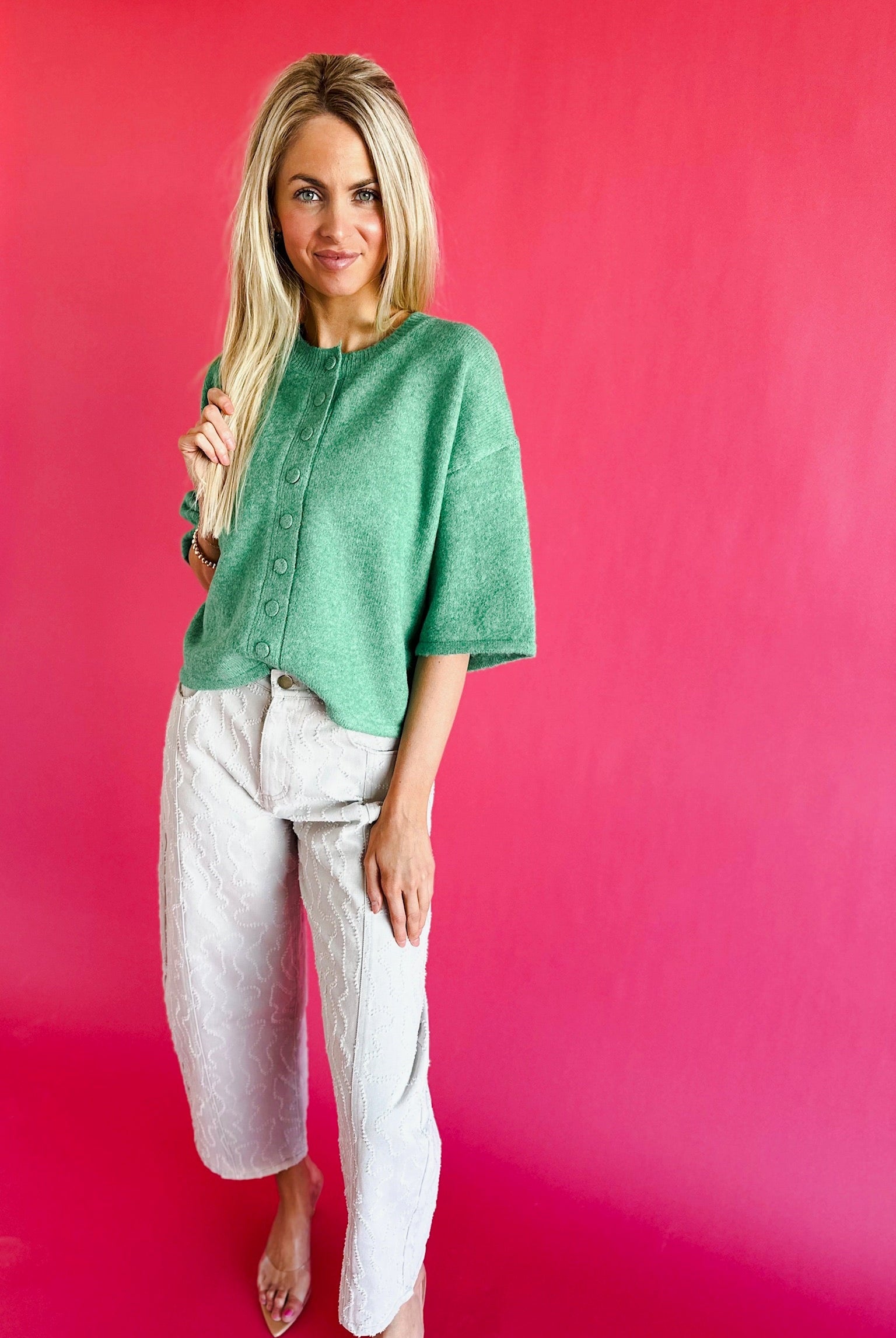 Woman wearing a green short sleeve knit sweater cardigan with button front, paired with light grey textured pants.