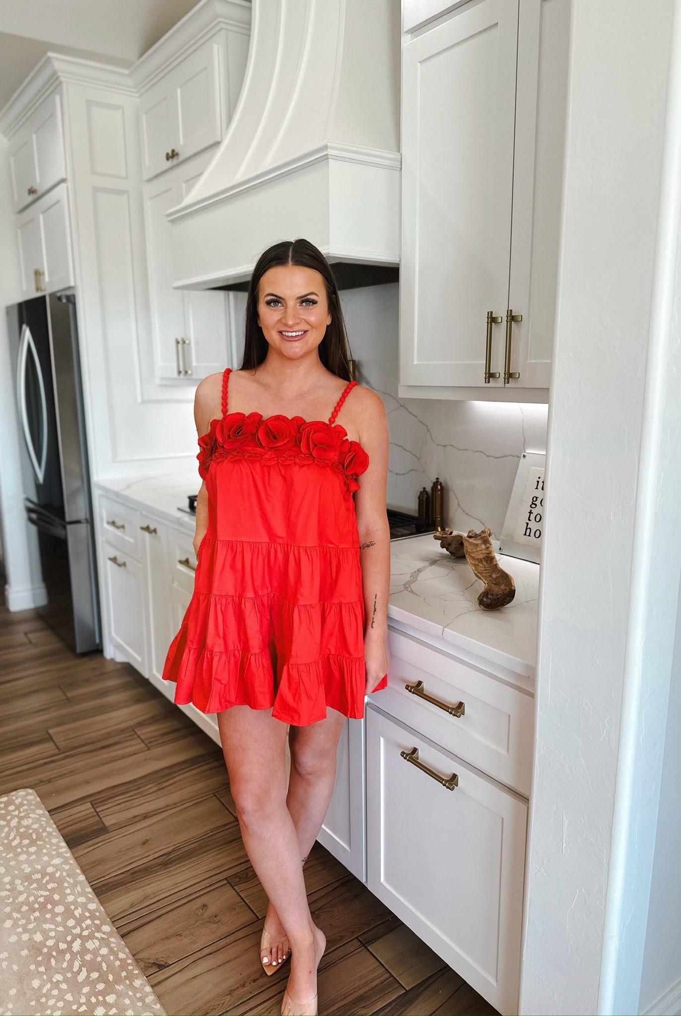 Woman in a red mini dress with floral straps and tiered skirt standing in a modern white kitchen.