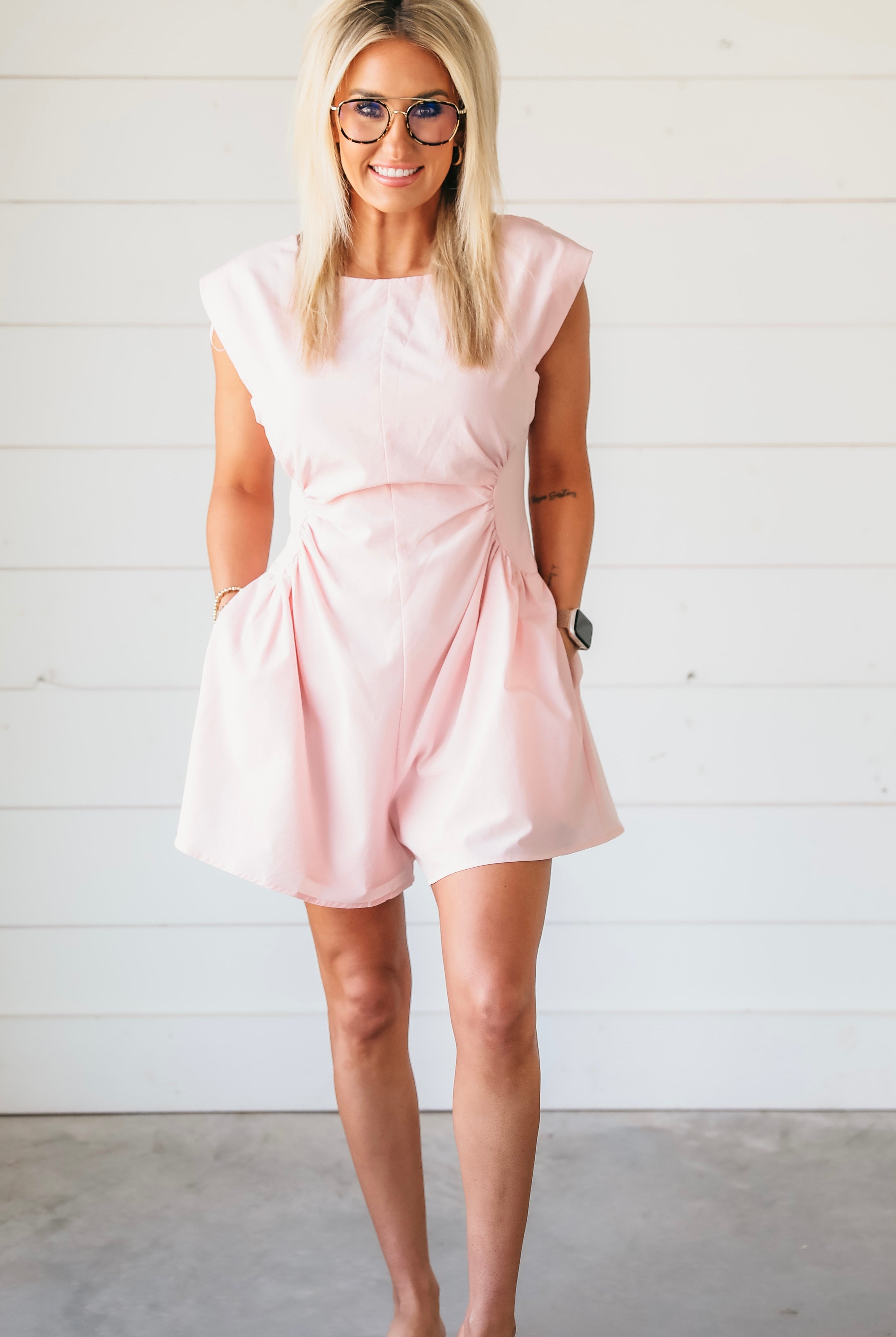 Woman wearing a powder pink romper with gathered waist detail, cap sleeves, and pockets. She has blonde hair, glasses, and
