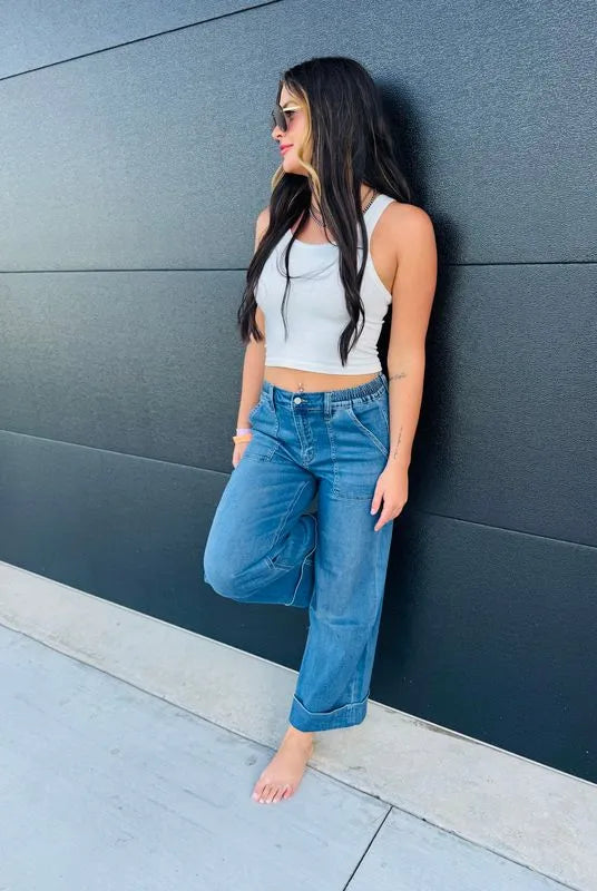 Woman wearing Vervet high rise double cuffed baggy wide leg blue jeans and a white crop top, leaning against a black wall.