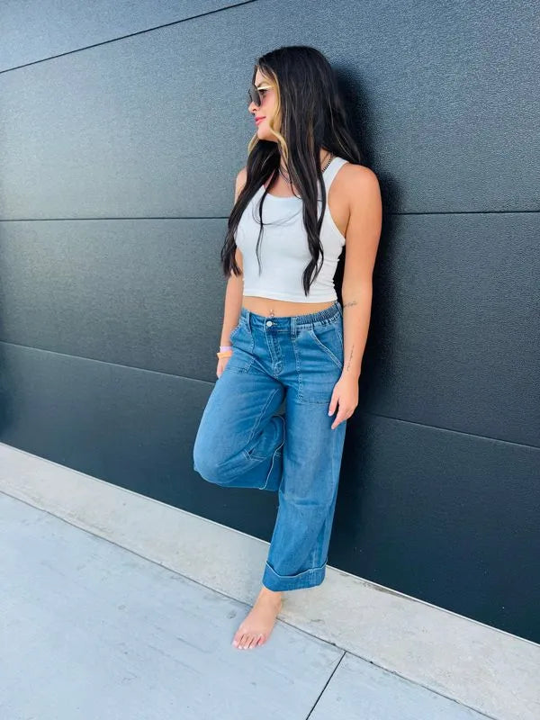 Woman wearing Vervet high rise double cuffed baggy wide leg blue jeans and a white crop top, leaning against a black wall.