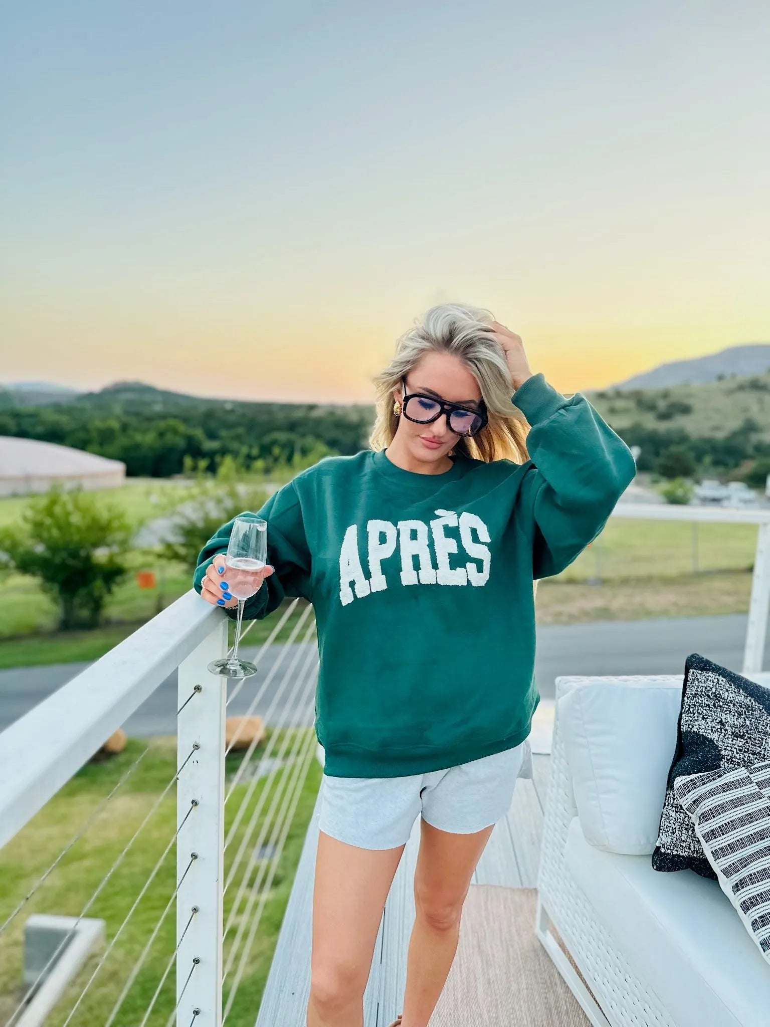Hunter green sweatshirt with white "APRÈS" graphic, worn by a woman with blonde hair and glasses, holding a champagne flute.