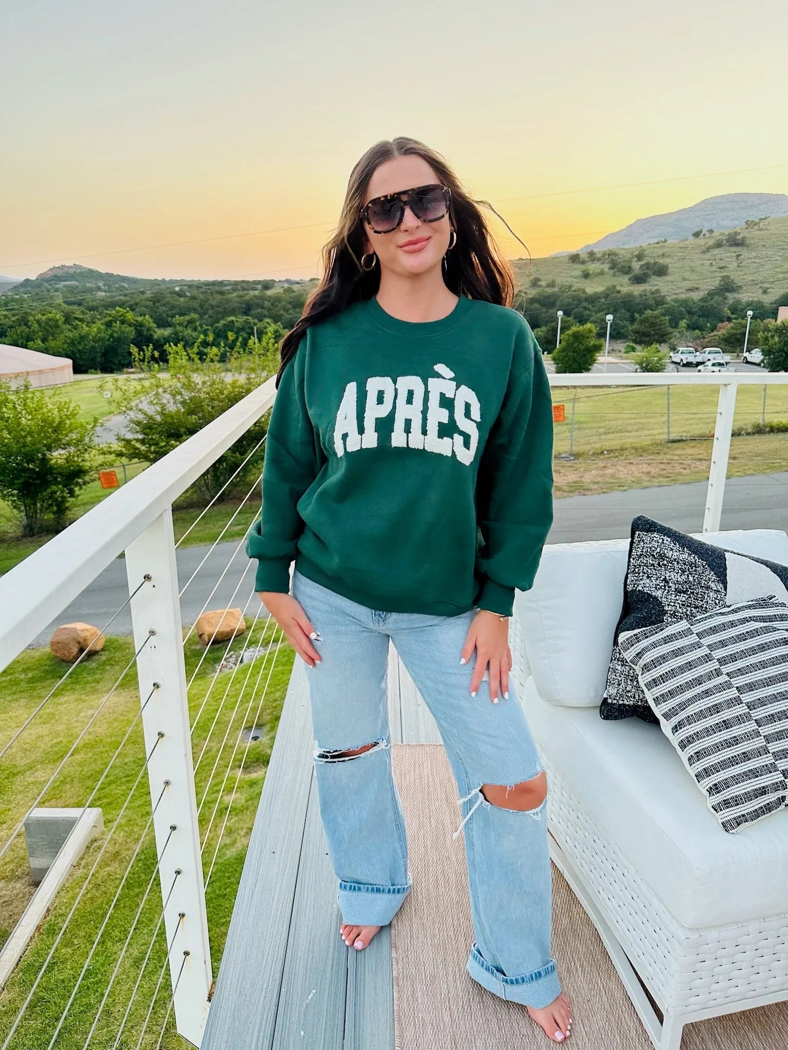 Hunter Green "APRÈS" sweatshirt on a woman wearing light wash ripped jeans, sunglasses, and hoop earrings.