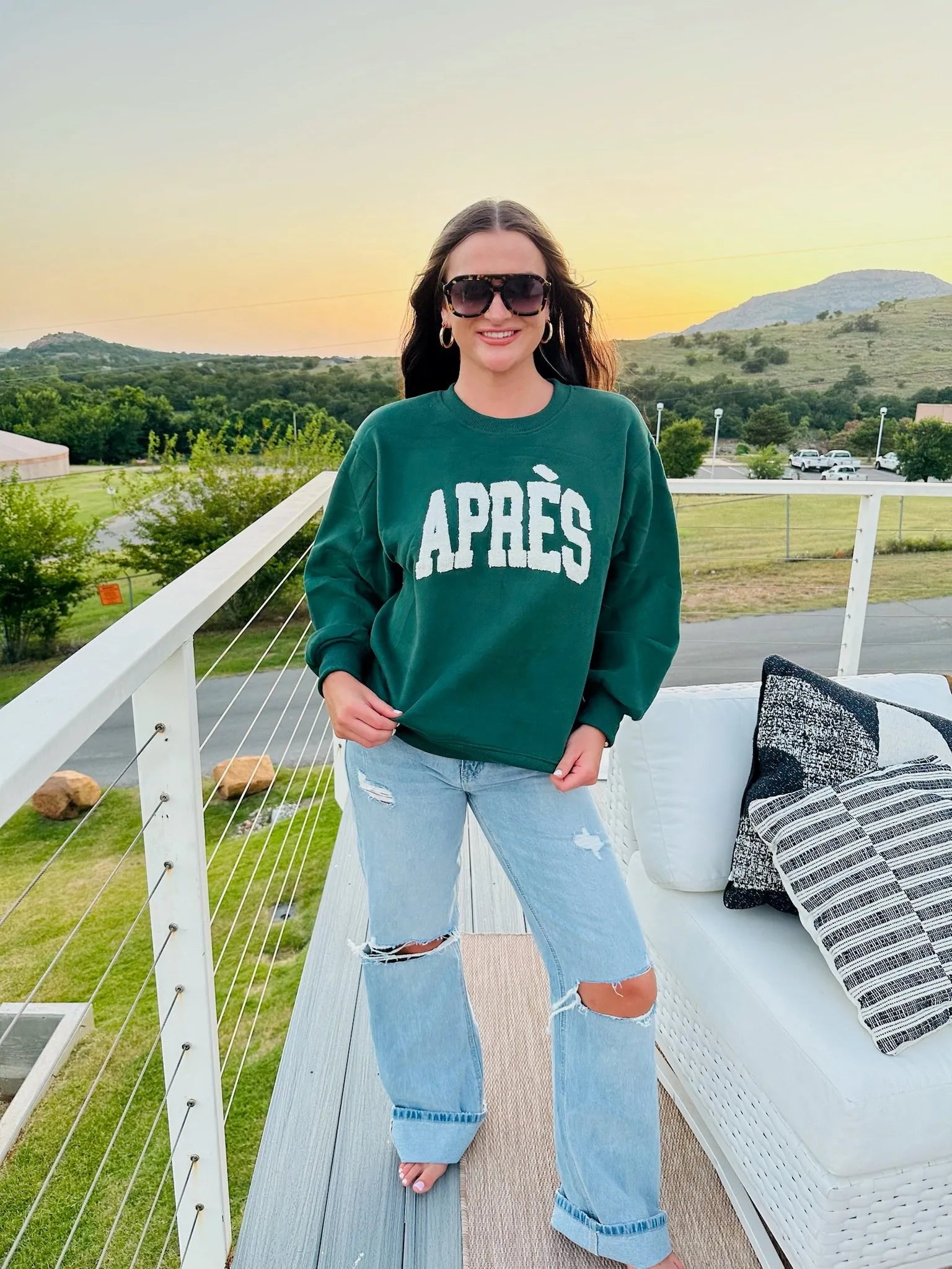 Hunter green crewneck sweatshirt with white "APRÈS" graphic, worn by a woman with sunglasses and ripped jeans.