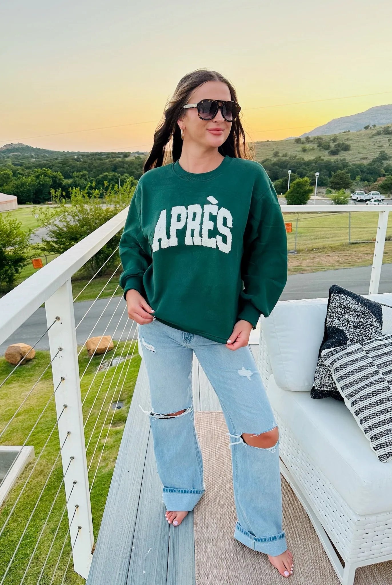 Woman wearing a hunter green sweatshirt with white "APRÈS" text and ripped light wash jeans.