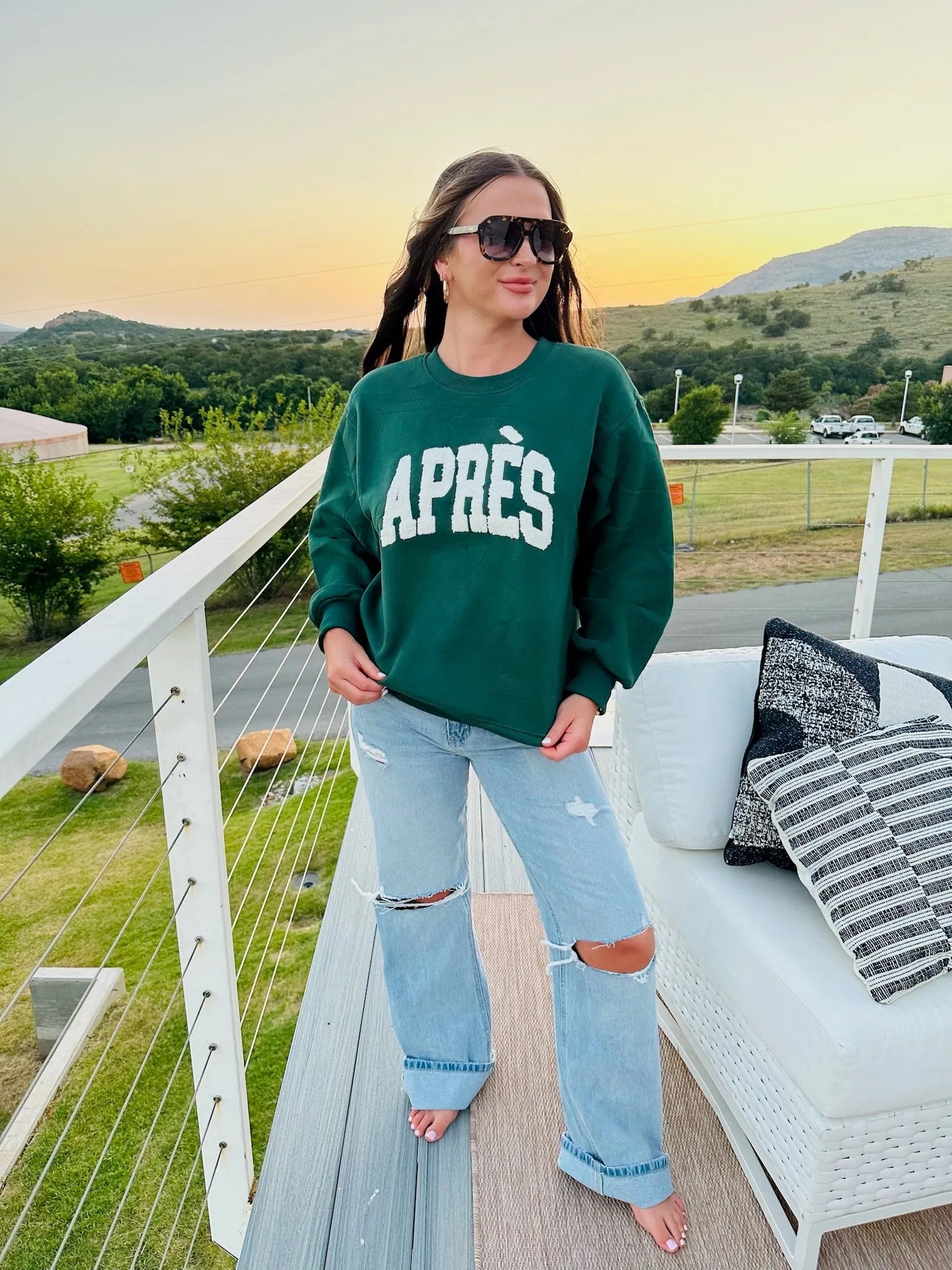 Woman wearing a hunter green sweatshirt with white "APRÈS" text and ripped light wash jeans.