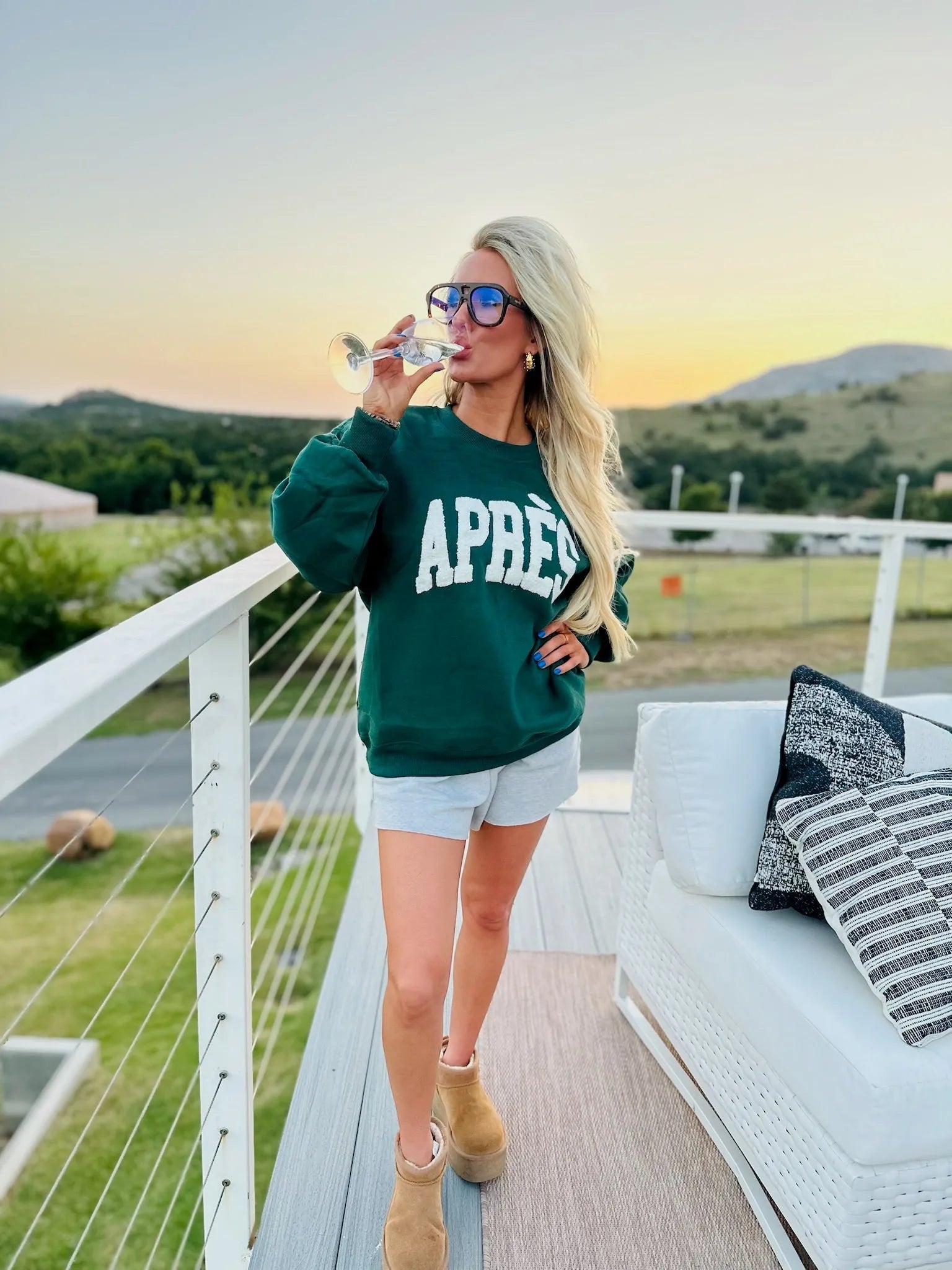 Hunter green sweatshirt with white "APR extEacute" graphic, worn by a woman with blonde hair, sunglasses, and grey shorts.