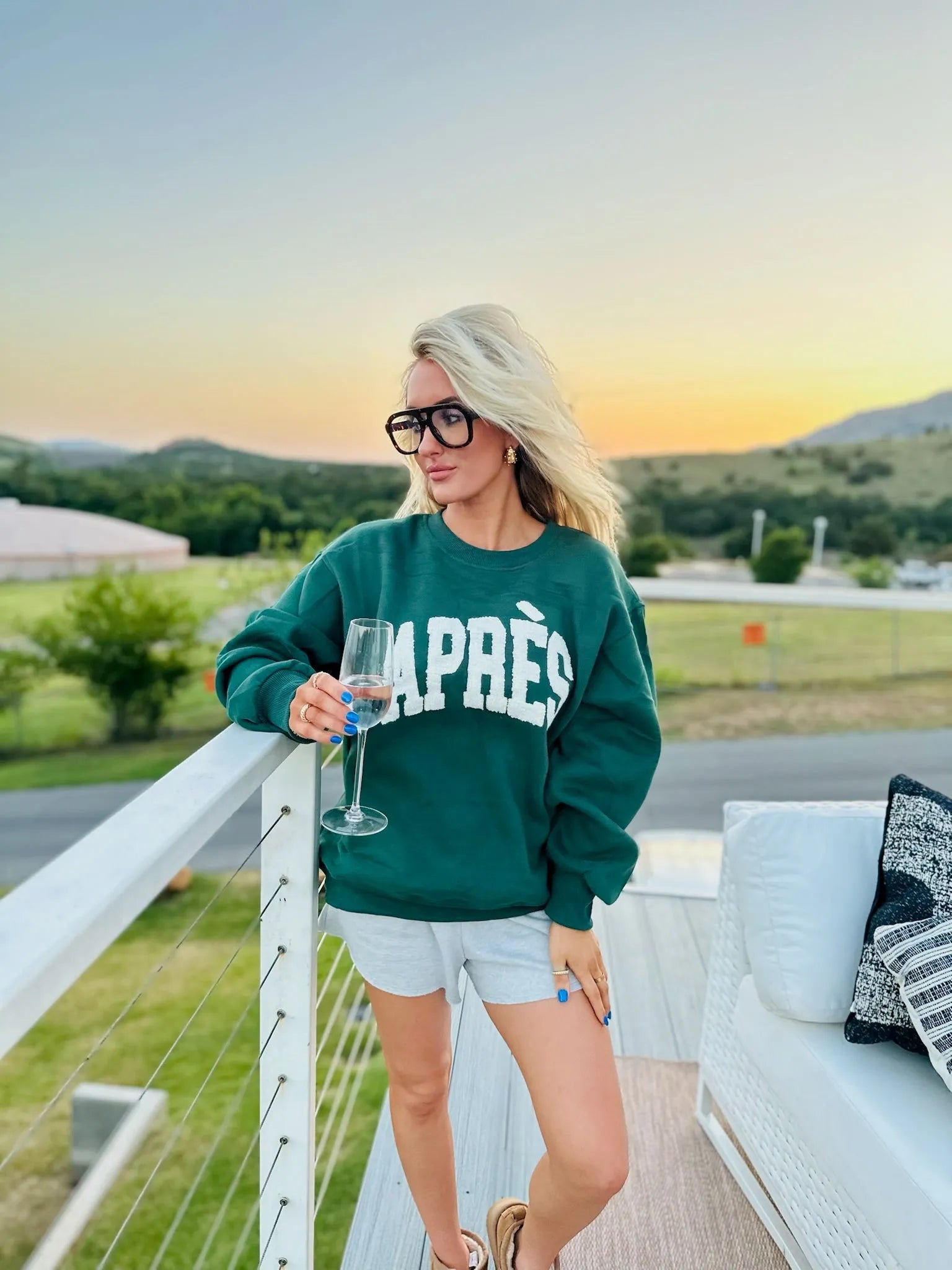 Hunter green sweatshirt with white "APRÈS" text. Woman holding wine glass, wearing glasses and grey shorts outdoors at