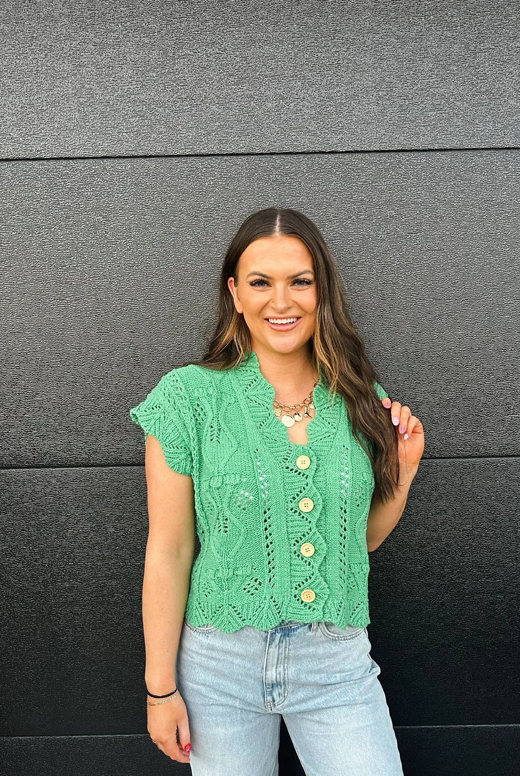 Green short-sleeve knit sweater with a lace pattern and wooden buttons. Model wears light wash jeans and a gold necklace.