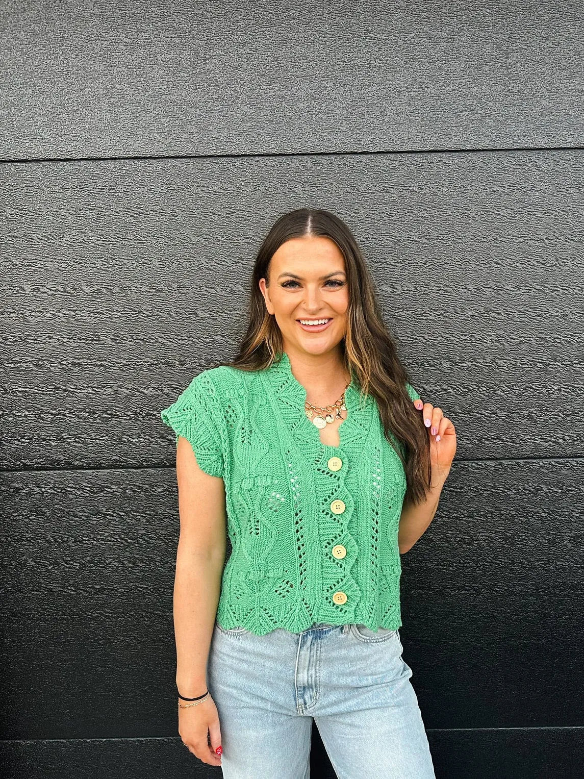 Green short-sleeve knit sweater with a lace pattern and wooden buttons. Model wears light wash jeans and a gold necklace.