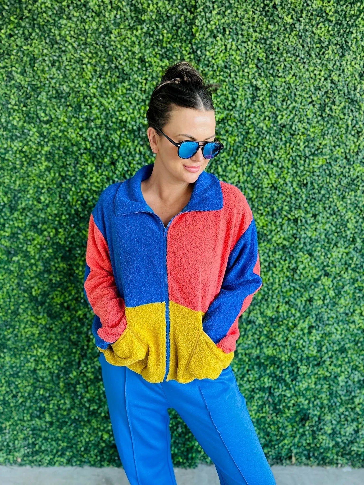 Woman wearing J.anna Sherpa Thunder color block zip-up jacket with coral, blue, and yellow panels. Blue sunglasses, blue