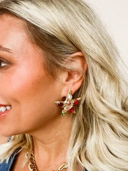 Gold star statement earrings with red, green, and white stones. Visible on a woman with blonde hair.