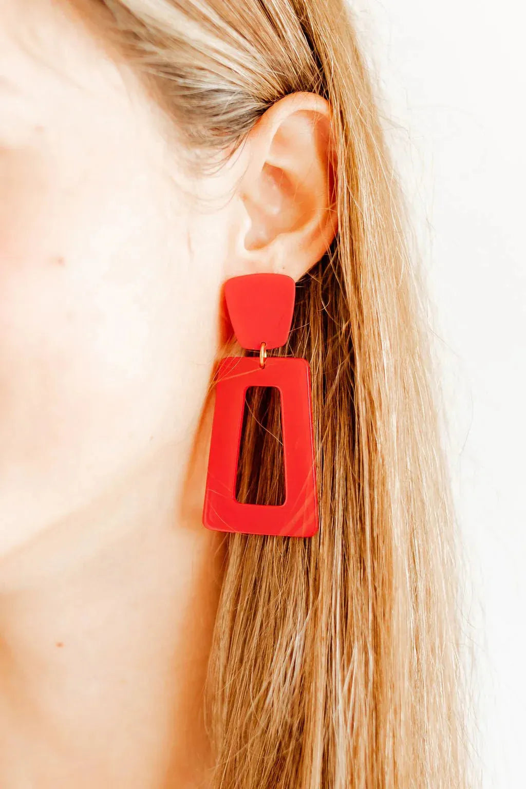 Red geometric statement drop earrings with a rectangular cutout, hanging from a woman's ear.