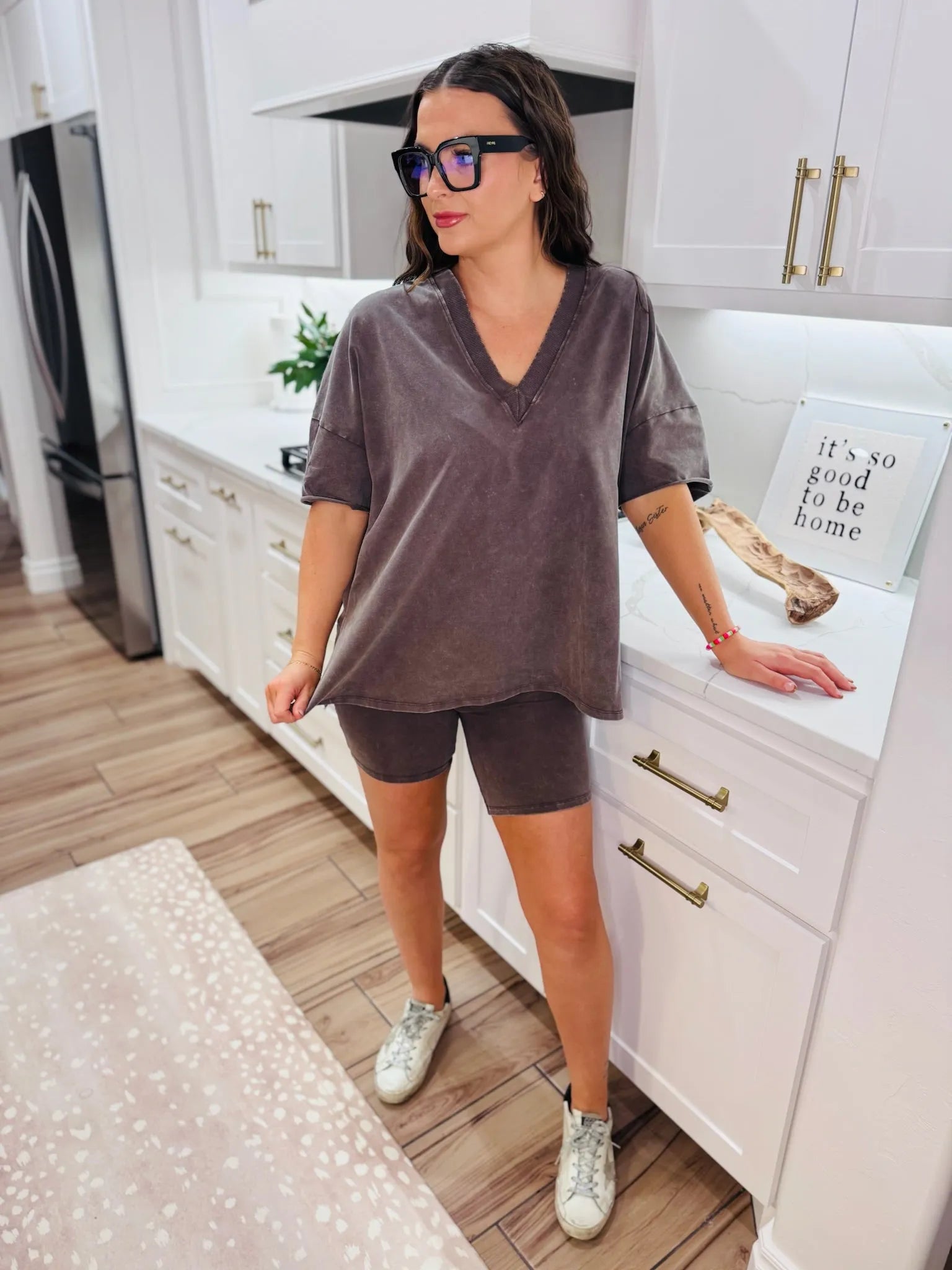 Woman wearing a dark grey v-neck t-shirt and matching dark grey biker shorts, paired with white sneakers. She has on large