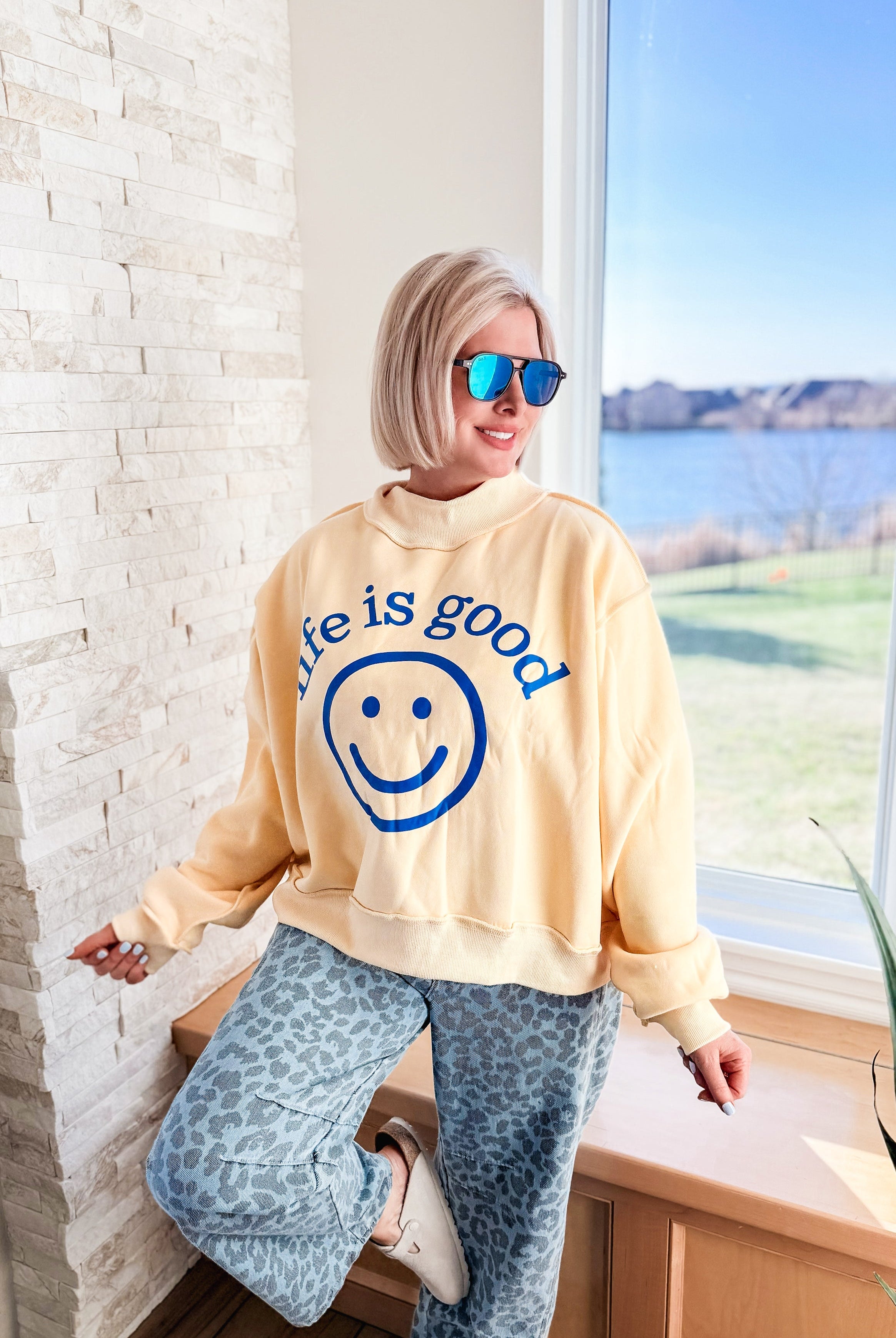 Woman wearing a yellow "Life is Good" graphic crop sweatshirt with a mock neck and blue leopard print pants.
