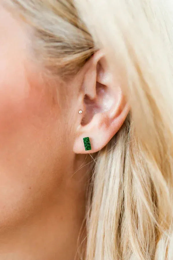 Close up of a woman's ear wearing small, rectangular green sparkly stud earrings. For sensitive ears.