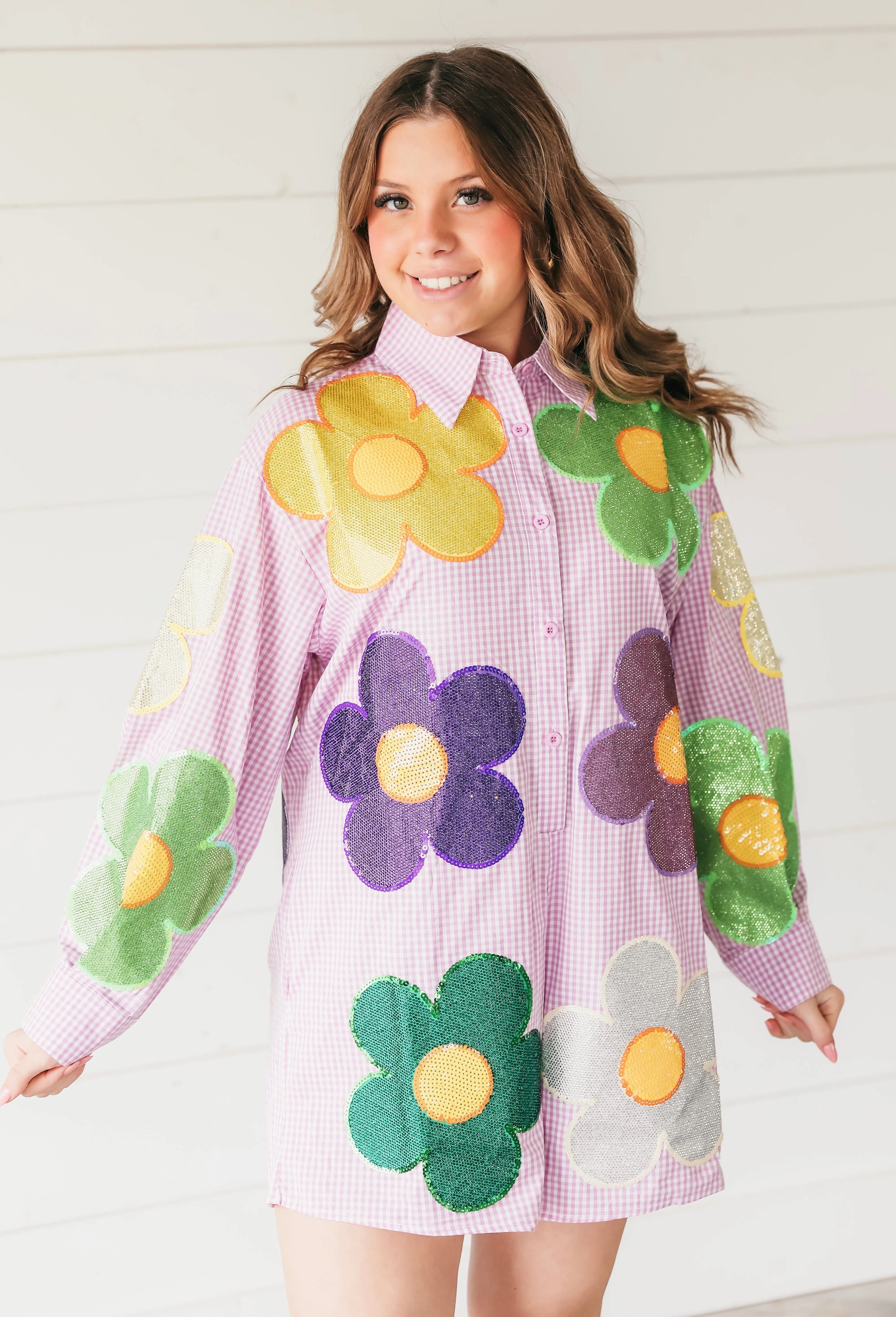 A woman wears a pink gingham romper adorned with large, colorful sequin flowers in yellow, green, purple, and silver.