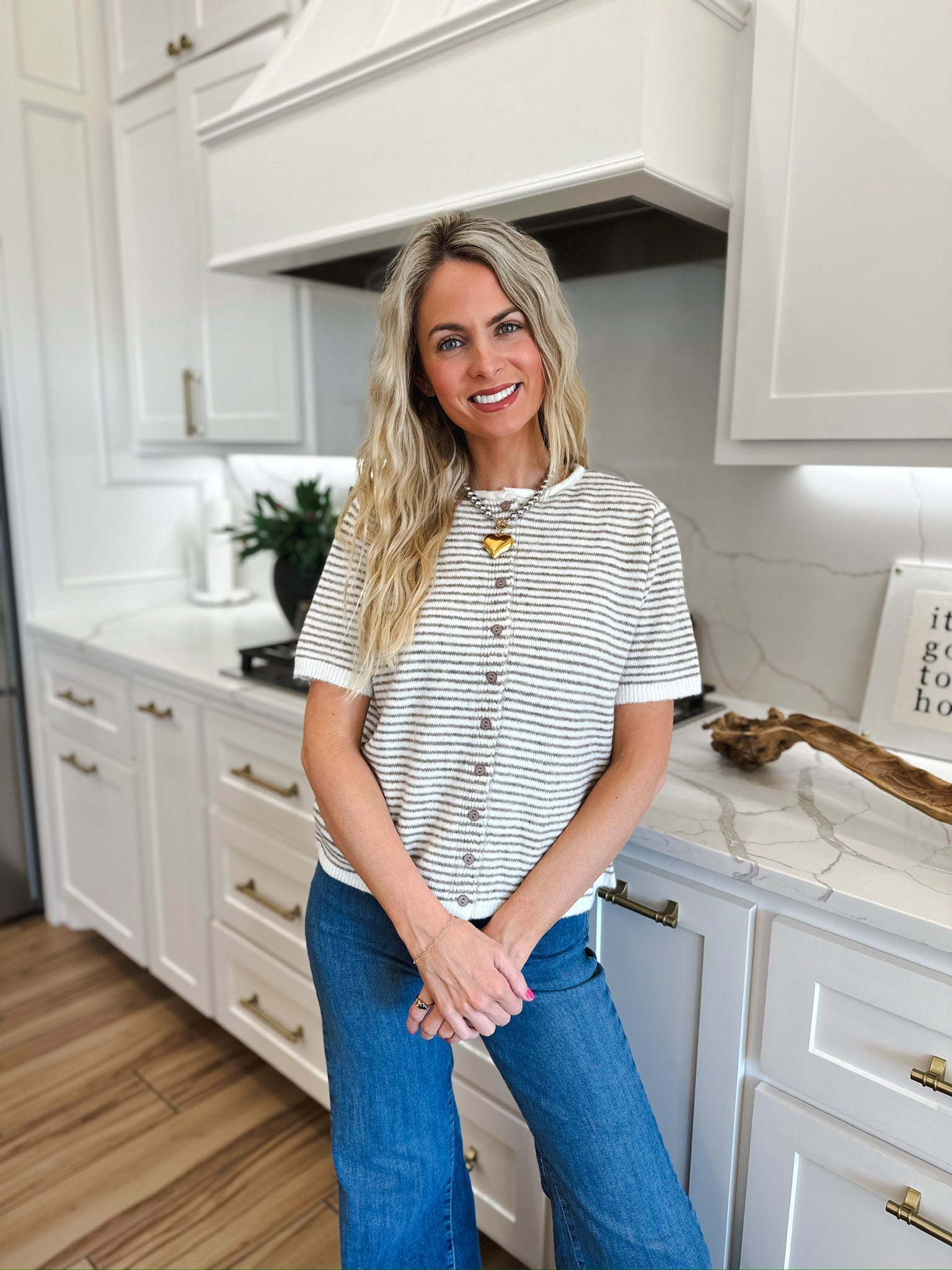 Woman wearing a cream and tan striped knit button-down short-sleeve top with a gold heart necklace and blue jeans.