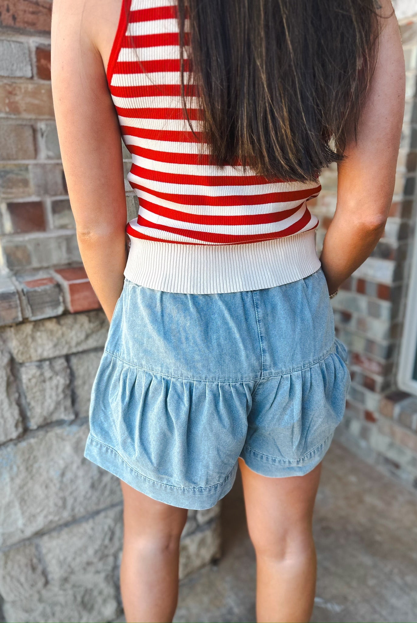 Back view of a woman wearing light blue mineral washed chambray shorts with a ruffled hem and a red and white striped tank