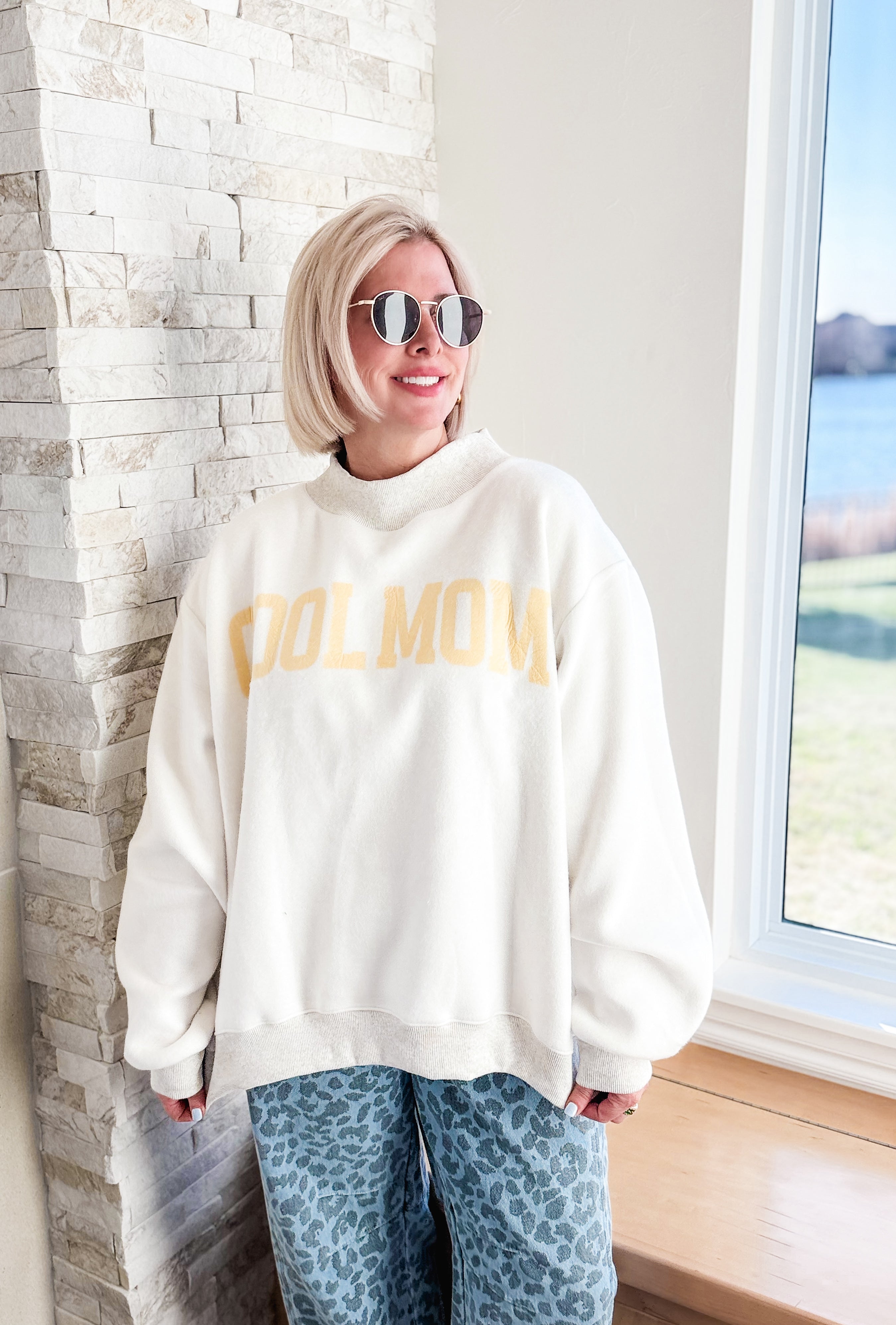 Woman wearing a cream "COOL MOM" graphic sweatshirt with a mock neck and light grey ribbed hem. She has on blue leopard