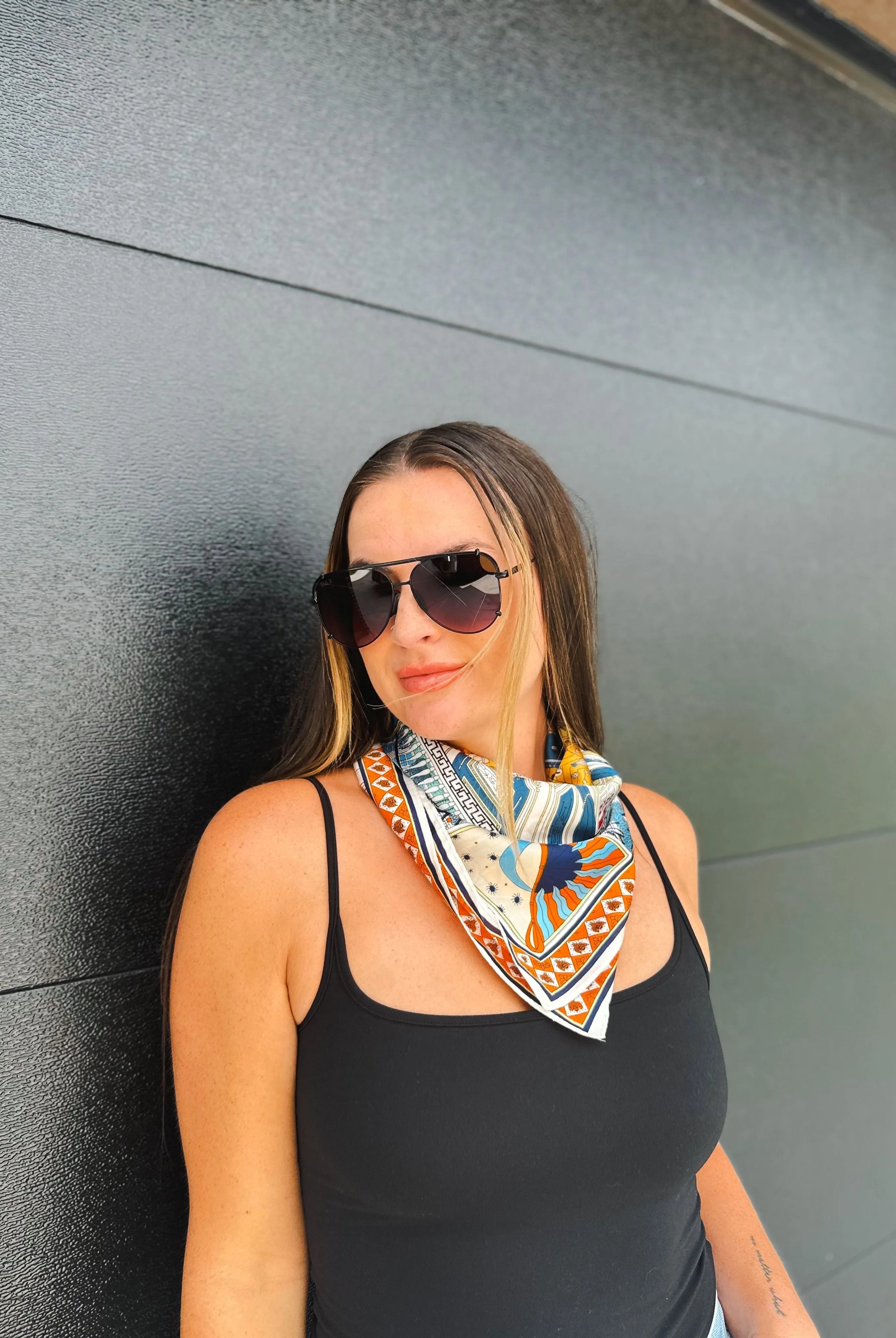 Woman wearing a black tank top and aviator sunglasses, with a colorful neck scarf featuring a sun and geometric print.