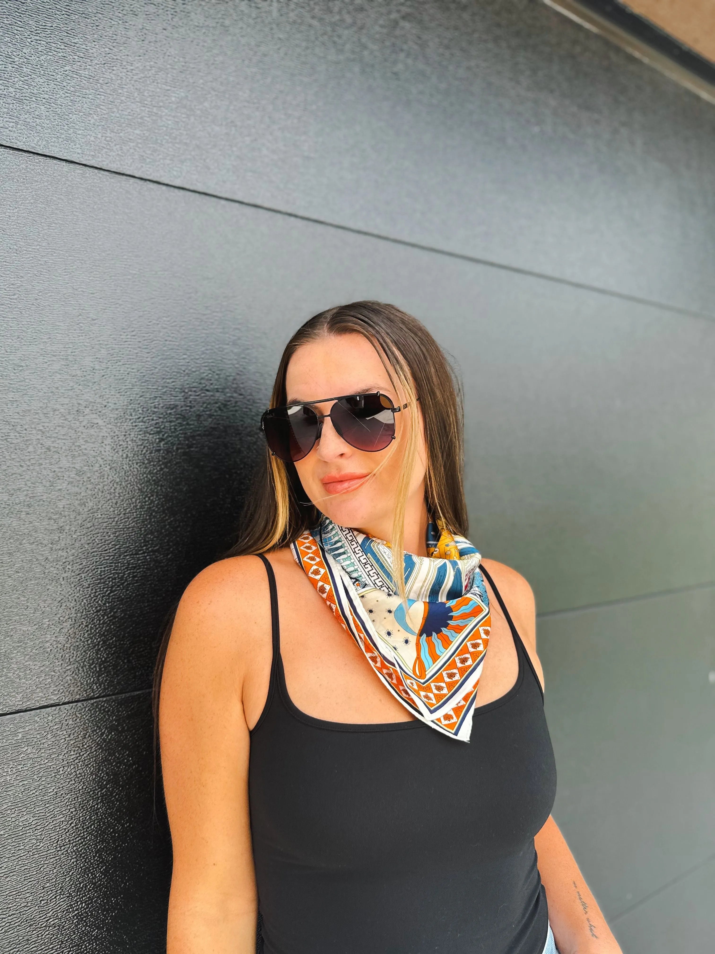 Woman wearing a black tank top and aviator sunglasses, with a colorful neck scarf featuring a sun and geometric print.