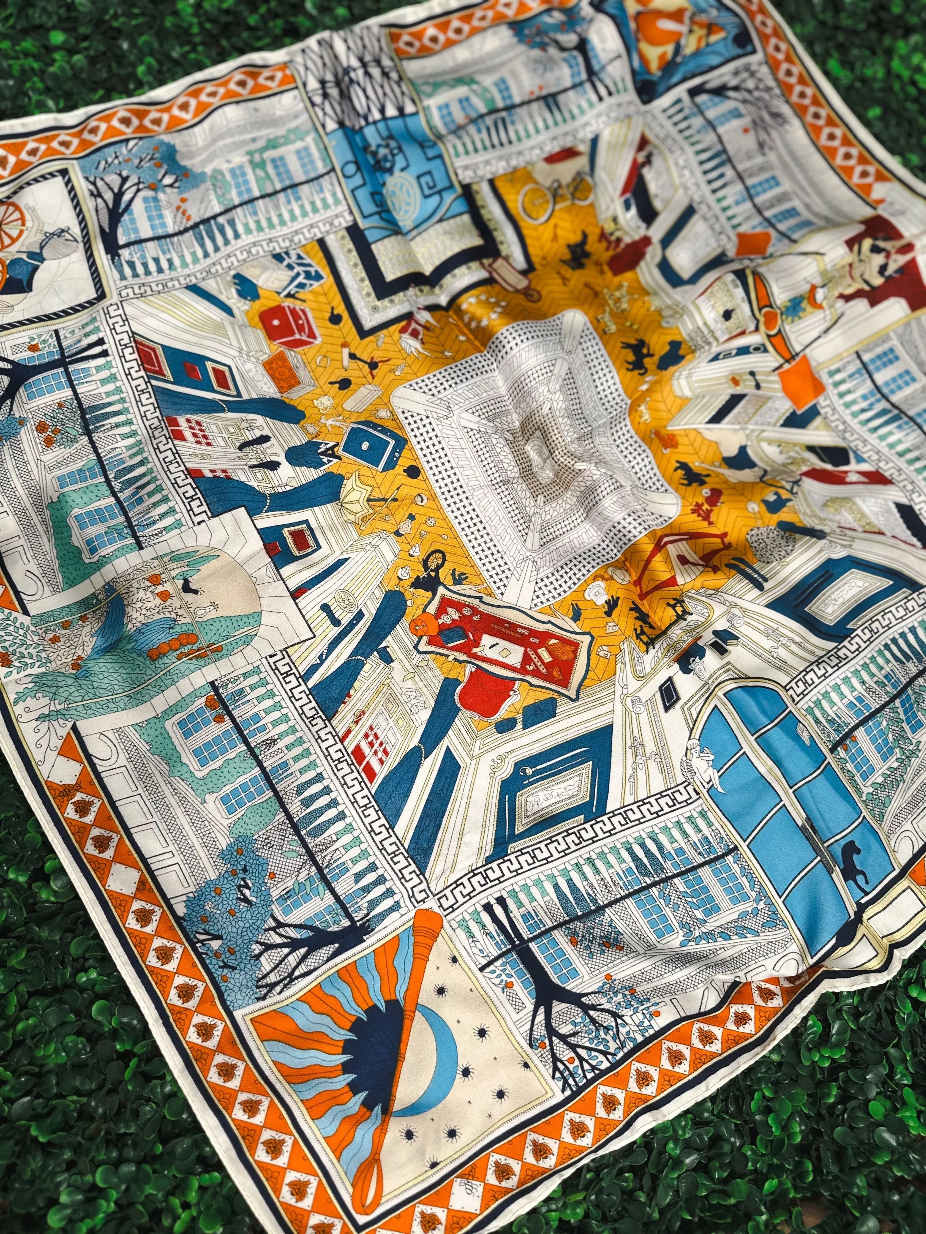 Colorful silk scarf with intricate pattern featuring architectural scenes, trees, sun, moon, and playful figures. Colors