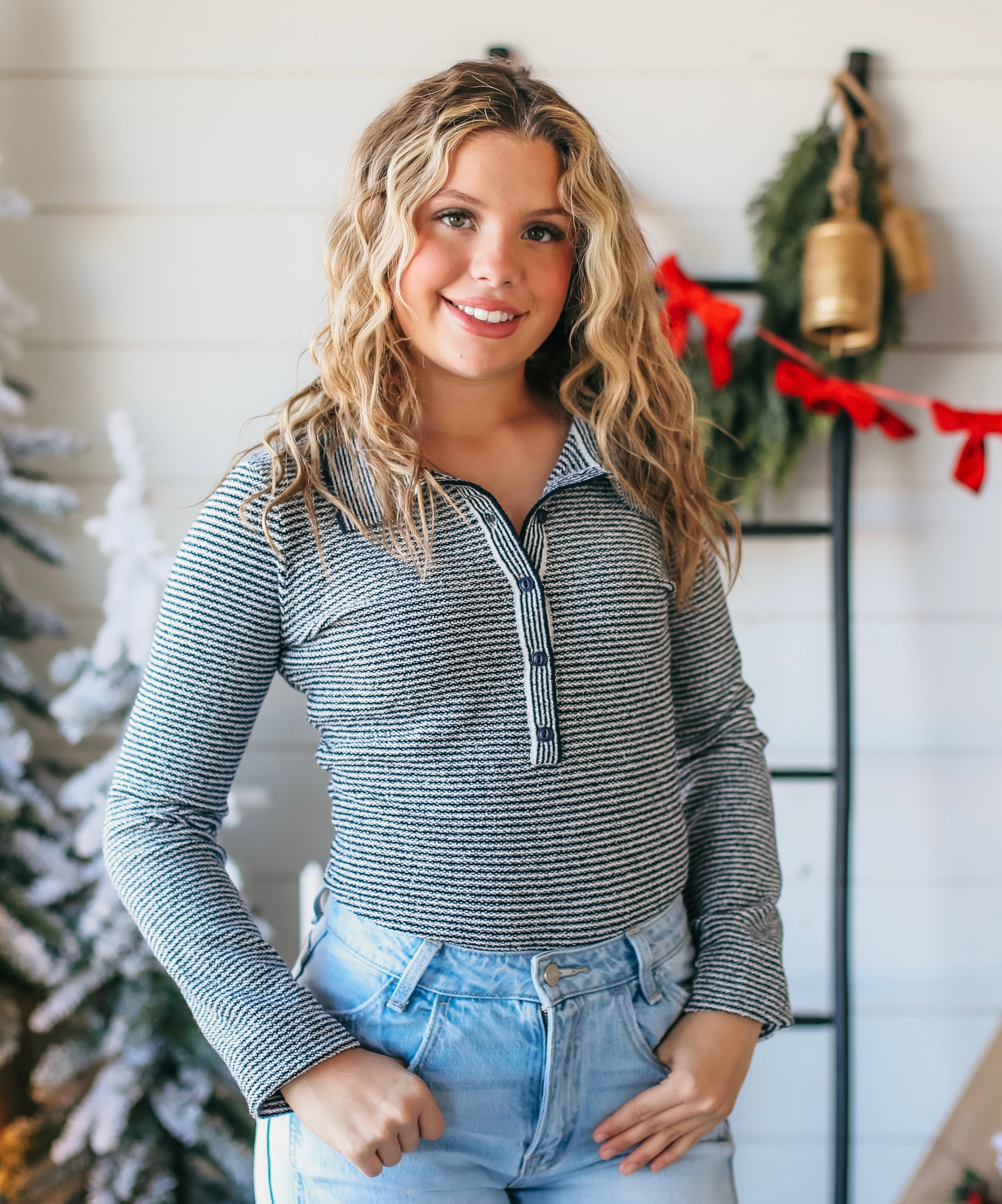 A young woman with blonde curly hair smiles as she poses in a navy and white striped collared long-sleeve bodysuit and