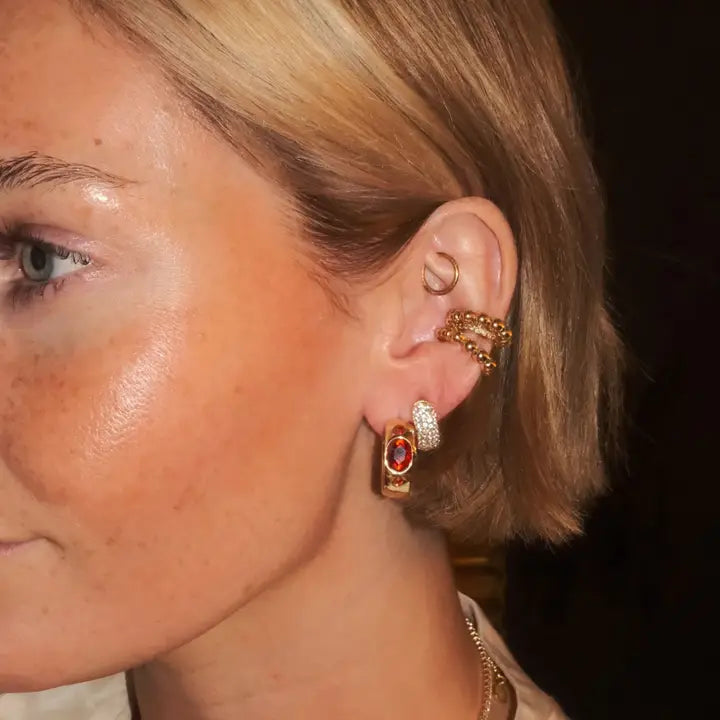 Gold hoop earrings with red oval CZ stones and pave CZ accents. Additional gold ear cuffs visible.