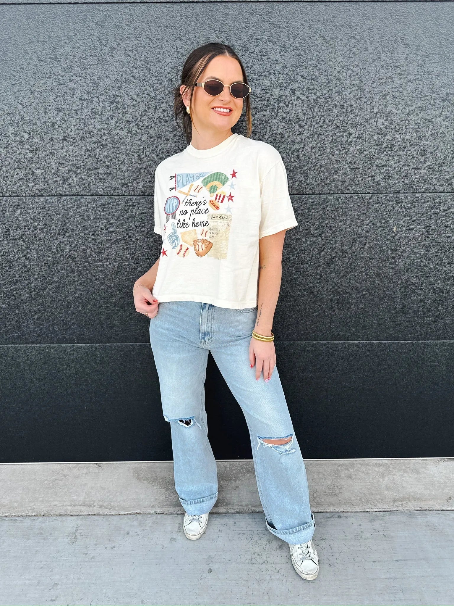 Cream graphic tee with baseball theme "There's no place like home" print, worn by a woman with light wash ripped jeans.