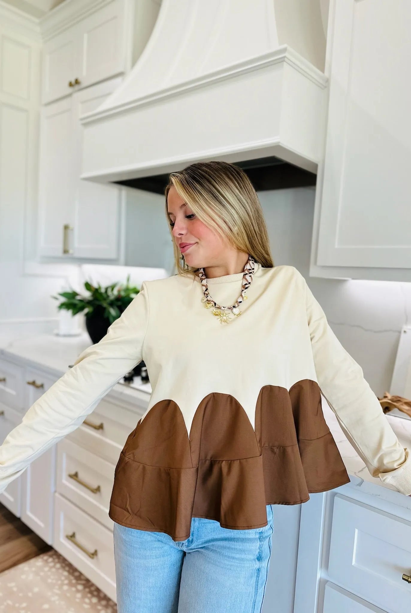 Woman wearing an oatmeal brown mixed media knit top with long sleeves and a ruffled hem, paired with light wash jeans and a
