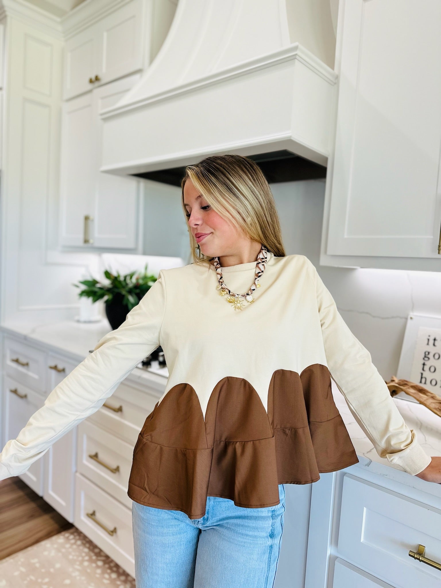 Woman wearing an oatmeal brown mixed media knit top with long sleeves and a ruffled hem, paired with light wash jeans and a