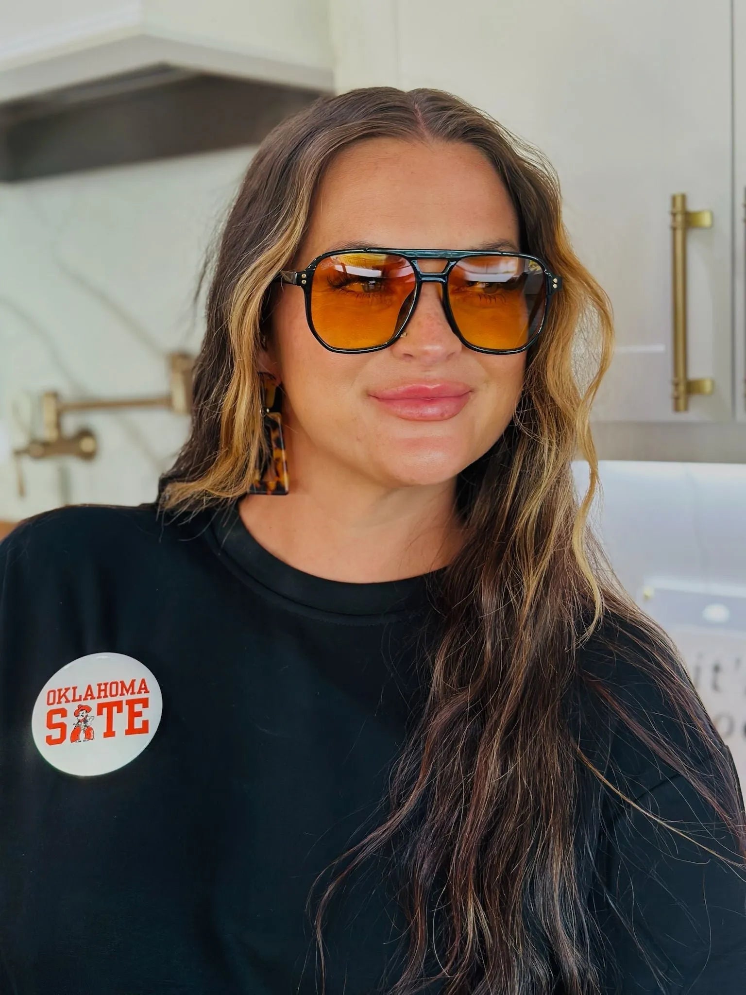 Round "Oklahoma State" button with mascot on black shirt, worn by a woman with large orange sunglasses and leopard print